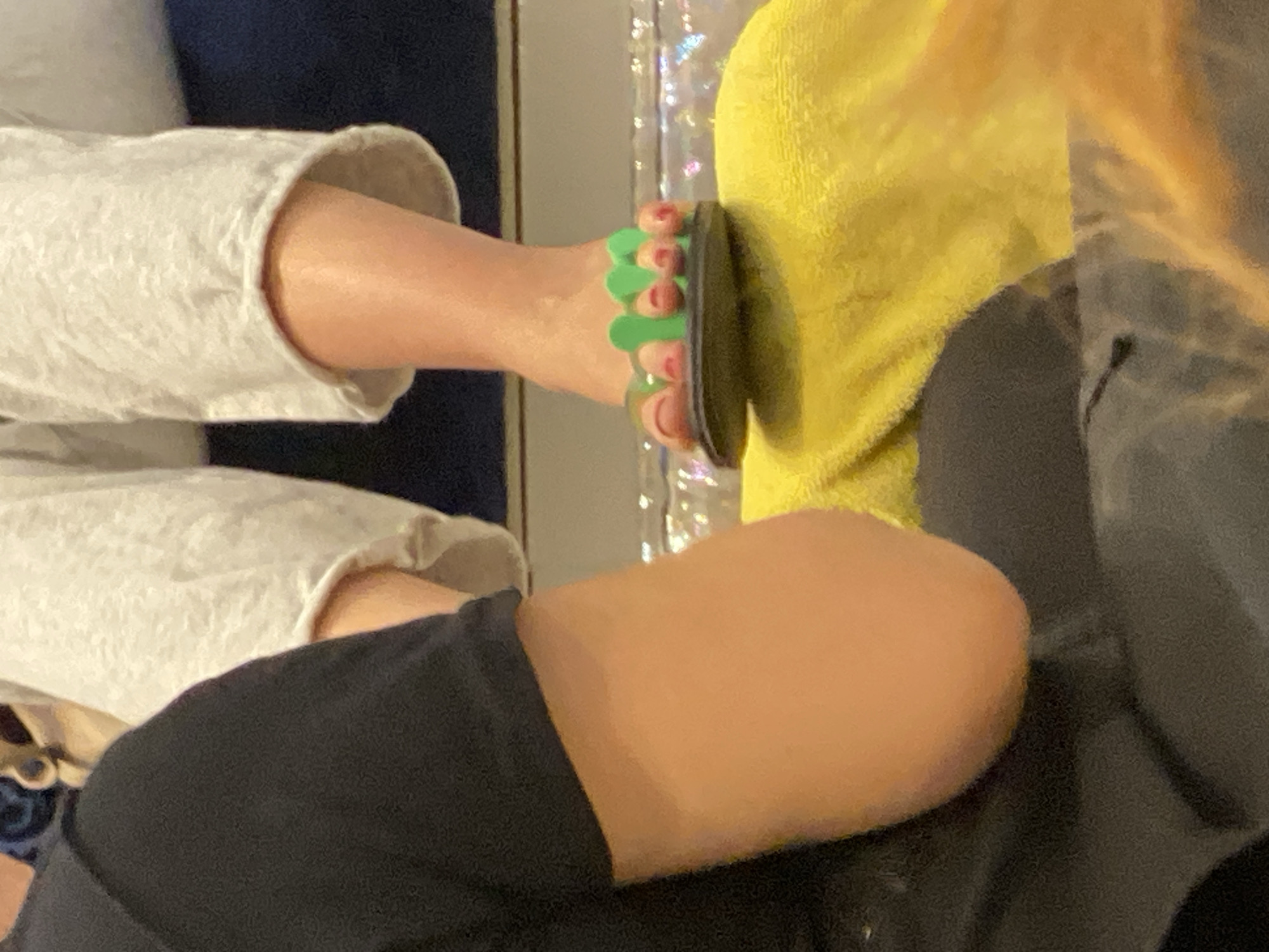 The image shows a person's foot wearing a green flip-flop. The person is wearing light-colored, loose-fitting pants. Part of another person's leg is visible, wearing dark-colored pants. A yellow towel or blanket is also visible. There is a shimmery, reflective surface in the background. There is no evidence of the image having been taken in New York City, United States of America.