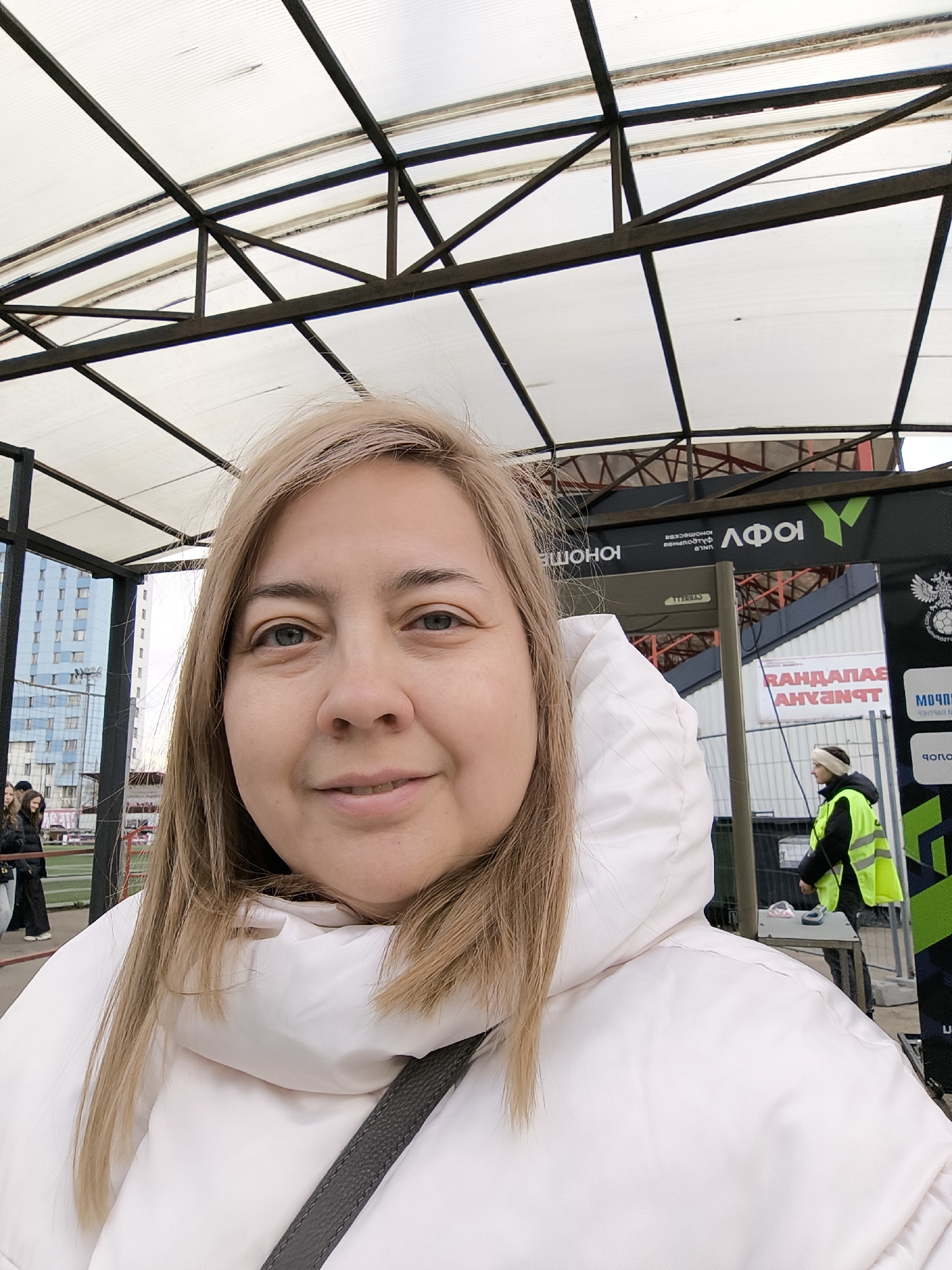 The image is a selfie of a fair-skinned, middle-aged woman with blonde hair and blue eyes, smiling at the camera. She is wearing a white puffer jacket with a large hood and a black strap across her body.
The background shows an outdoor setting, likely in the city of Khimki, Russia. Overhead is a metal structure with a white, translucent roof. Behind the woman and to the right is an entrance area with signage in Russian. A person wearing a high-visibility yellow vest is standing near a table, possibly a security checkpoint. In the distance on the left is a building, a green sports field, and other people.