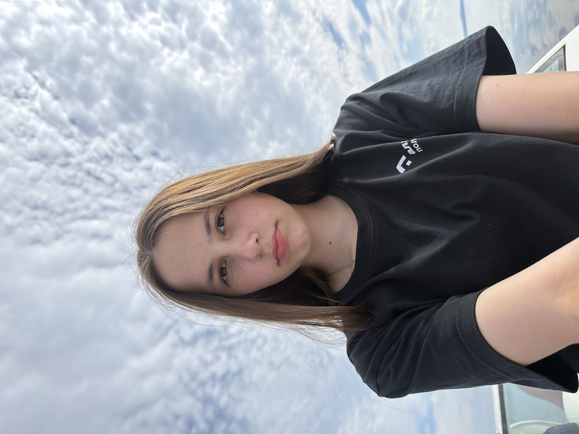 A young person with light brown hair and freckles, wearing a black t-shirt, is captured in a close-up, waist-up shot. Their gaze is directed forward, and their lips are slightly parted. The background consists of a cloudy sky. There is a logo on the t-shirt on the person's right side, which appears to be in Cyrillic script. The scene is outdoors, with a body of water and a light-colored railing visible at the bottom of the frame, suggesting the person is on a boat or near a body of water. It is not possible to identify the location in the city of Chaykovsky, Russia, from the provided image.