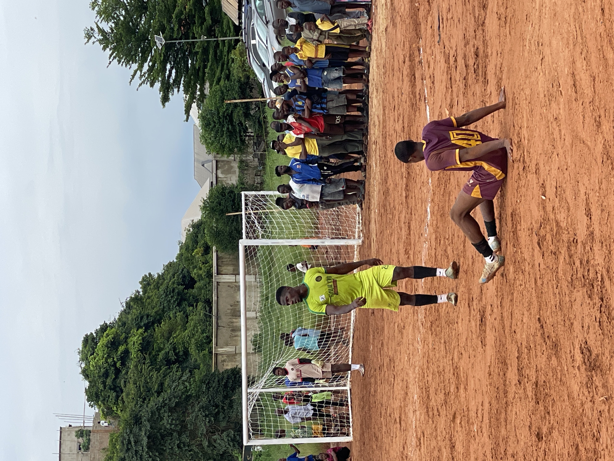 In an outdoor scene on a red dirt soccer field in Unknown, Unknown, two soccer players are the central focus. One player, wearing a bright green jersey and shorts with black socks and white cleats, stands looking to his right. The other player, dressed in a burgundy and gold jersey with the number 40 on the back, sits on the ground with his back to the viewer, his right hand supporting him as he leans to his side. His left leg is bent, and his right leg is extended. He wears black socks and cleats.
Behind them, a soccer goal with a white net stands. A group of spectators, mostly men and some boys, are gathered behind the goal, watching the game. They are dressed in casual clothing. Some spectators are standing, while others are seated on the ground.
In the background, beyond the spectators and goal, there is a concrete wall and lush green trees. Further back, buildings and cars are visible, suggesting a semi-urban or suburban environment. The sky is overcast. The overall impression is a casual soccer match taking place in a somewhat rustic setting.