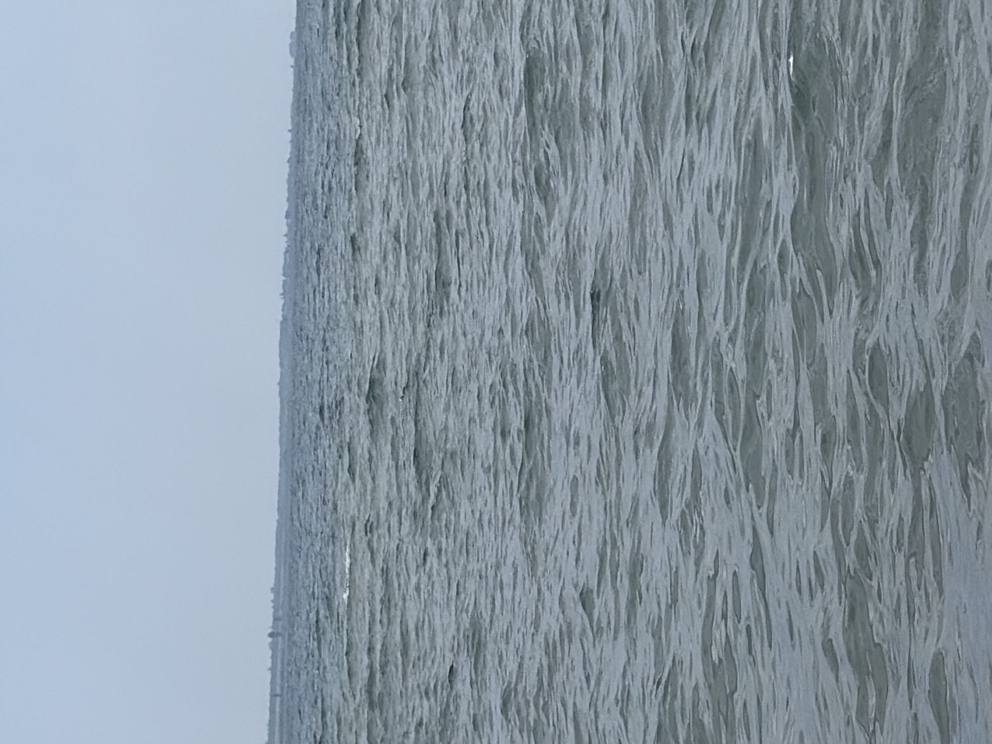 The image depicts a wide expanse of choppy, gray water under a pale, overcast sky. The horizon line is visible in the distance, where a faint, low-lying landmass with indistinct structures can be discerned. The water is covered in small, undulating waves, creating a textured surface. There are no people, animals, or prominent objects in the foreground or middle ground. The scene conveys a sense of open space and a muted, possibly cool, atmosphere.