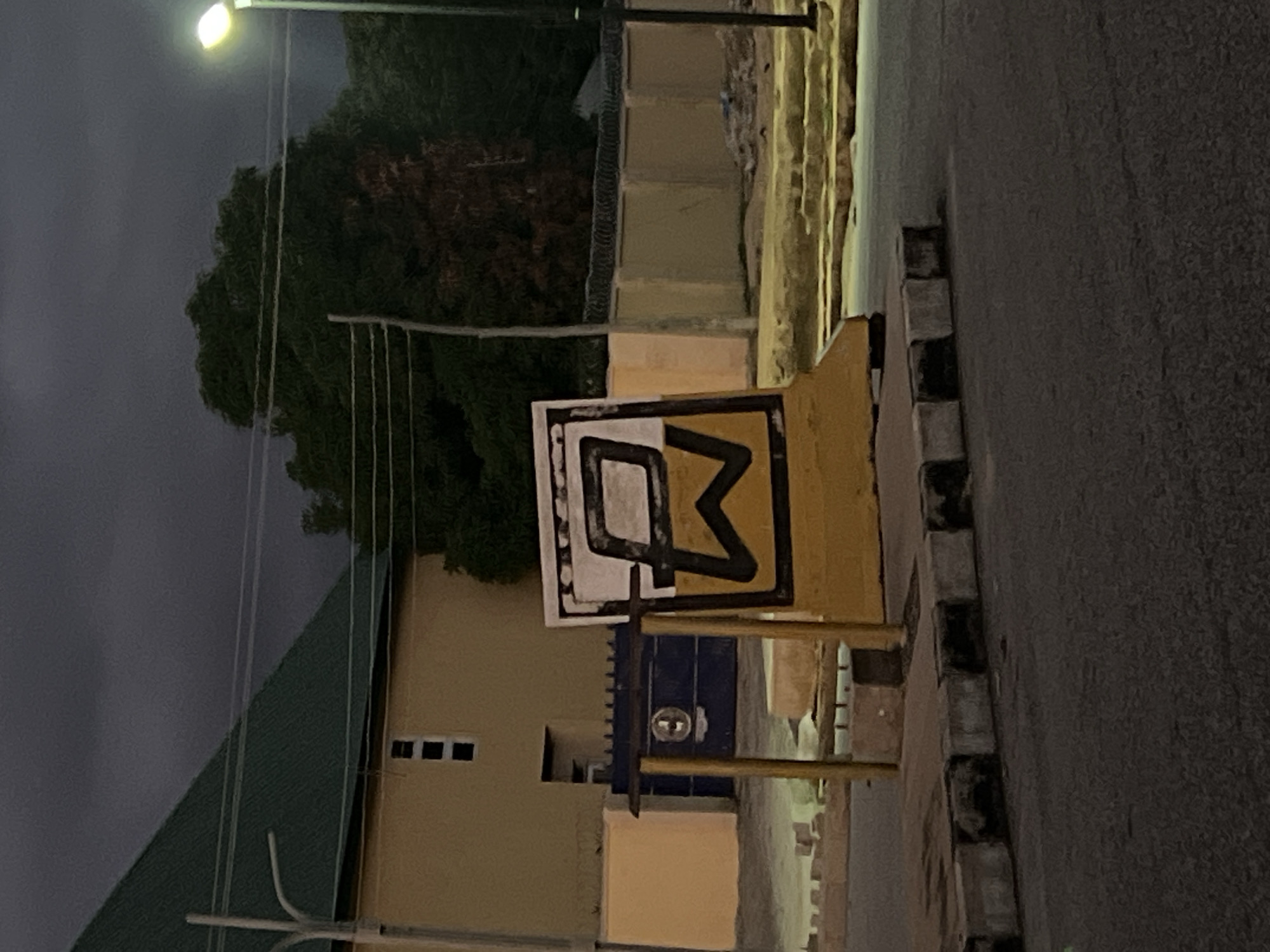 This image captures an outdoor street scene in Jalingo, Nigeria, under low light conditions, indicative of either late dusk or night, beneath a uniformly dark, overcast sky.
In the foreground, an asphalt road surface is visible, bordered by a concrete curb painted in alternating grey and black sections. A prominent yellow concrete barrier or base supports a rectangular sign. The sign itself features a white background encased by a thick black border, within which two stylized black capital letters are displayed: an upper 'P' with a distinct angular cut in its loop, and a lower 'W' with a sharp central peak, sharing a common vertical element.
Behind the sign, a light beige wall extends to the left. To the right of the sign's base, a dark blue metal gate is partially visible, featuring a circular emblem on its left panel. Beyond the gate, a building with a light beige facade and a dark green sloped roof is present. This building has three small, vertically aligned rectangular windows on its upper section. The beige wall continues to the right, topped with multiple strands of barbed wire. Behind this wall, dense, dark green tree foliage is visible.
On the far right, a tall street light pole with an illuminated, bright yellow-white lamp casts light into the scene. On the far left, another street light pole, without an active lamp, is also visible. Several overhead utility lines span horizontally across the upper midground and background, against the dark sky and above the trees. No individuals are present in the frame.