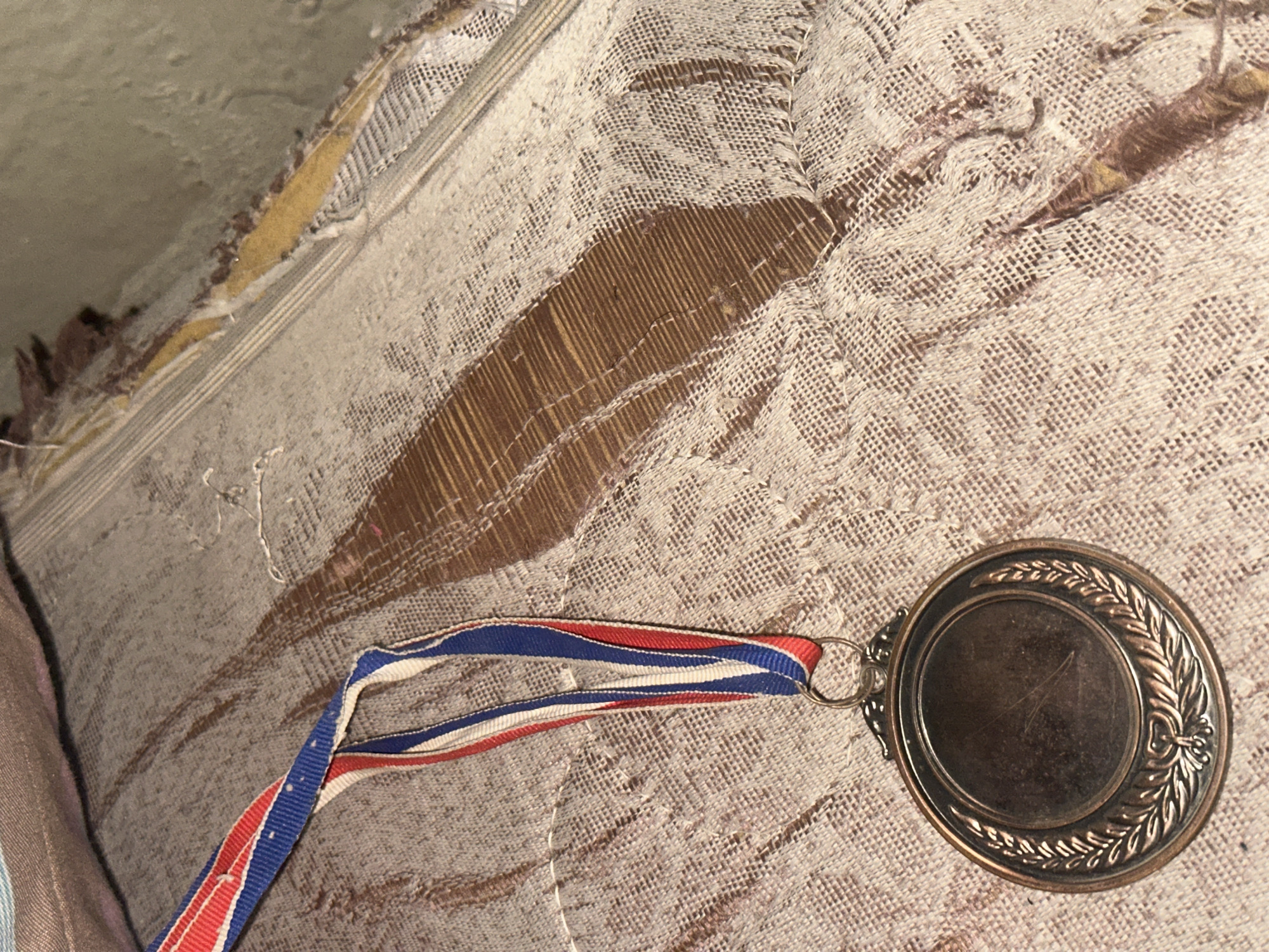 A close-up shot of a medal with a red, white, and blue ribbon hangs on a textured, woven surface. The medal is circular with a bronzed finish and features a laurel wreath design around the edge. The ribbon is attached to the medal by a ring. The background surface appears worn and damaged, with visible threads and a wooden or cardboard substructure showing through. The lighting is dim and casts shadows, creating a sense of depth and texture. The scene is devoid of any identifiable context related to a specific location in a city.