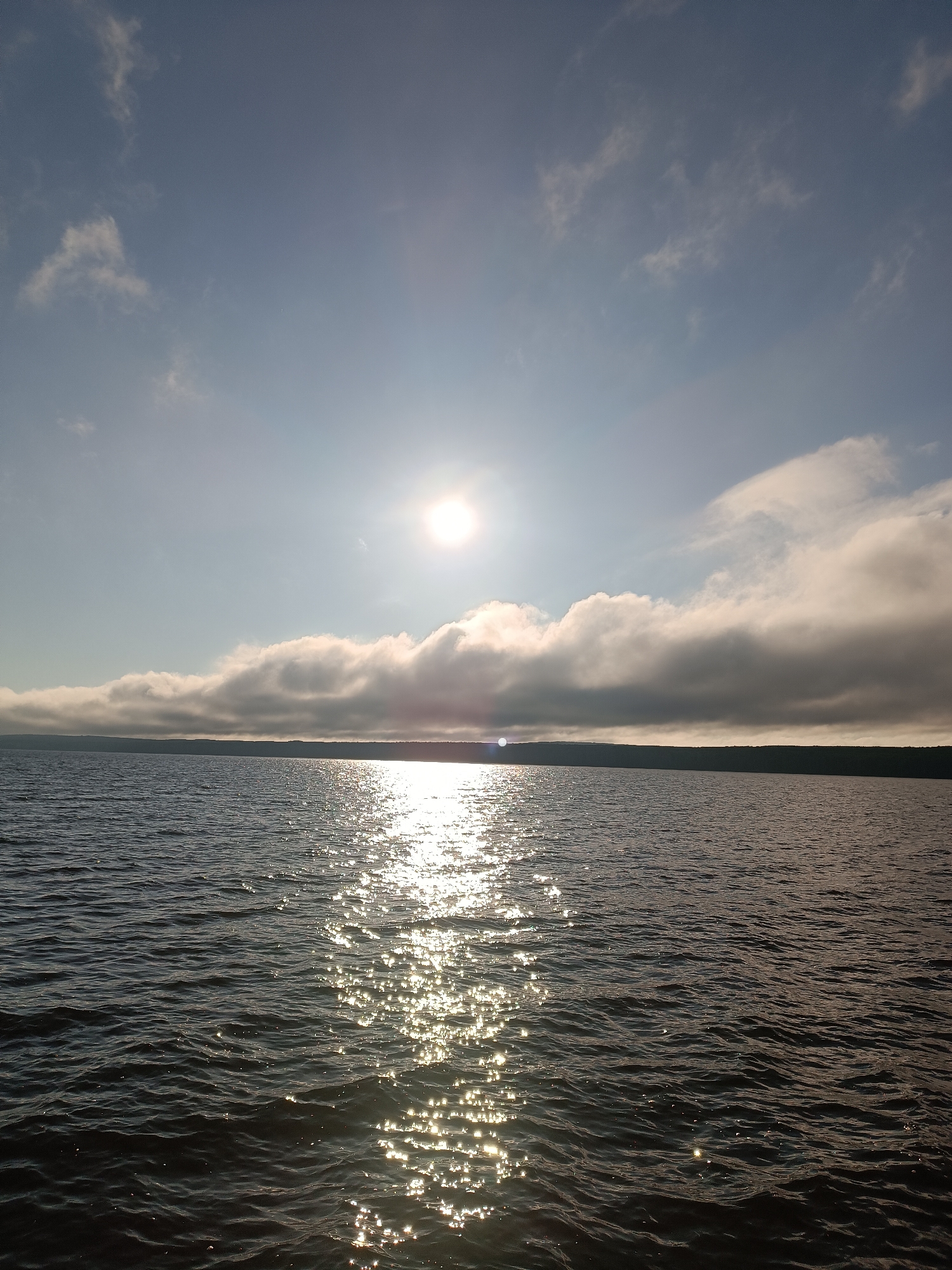 The image depicts a wide expanse of dark, rippling water, likely a lake or a large river, under a bright, hazy sun. The sun, located in the upper left quadrant of the image, casts a strong reflection across the water's surface, creating a shimmering path of light. A line of dark green trees forms a distant horizon across the width of the image. Above the trees, a bank of thick, white and grey clouds stretches horizontally, partially obscuring the lighter blue sky. The overall scene suggests an outdoor natural environment, possibly at dawn or dusk given the low angle of the sun and the atmospheric conditions. This scene could be located on the Kama River or one of its reservoirs near Chaikovsky, Russia, a city known for its proximity to large bodies of water and hydroelectric power generation. There are no people or specific objects clearly visible in the image.