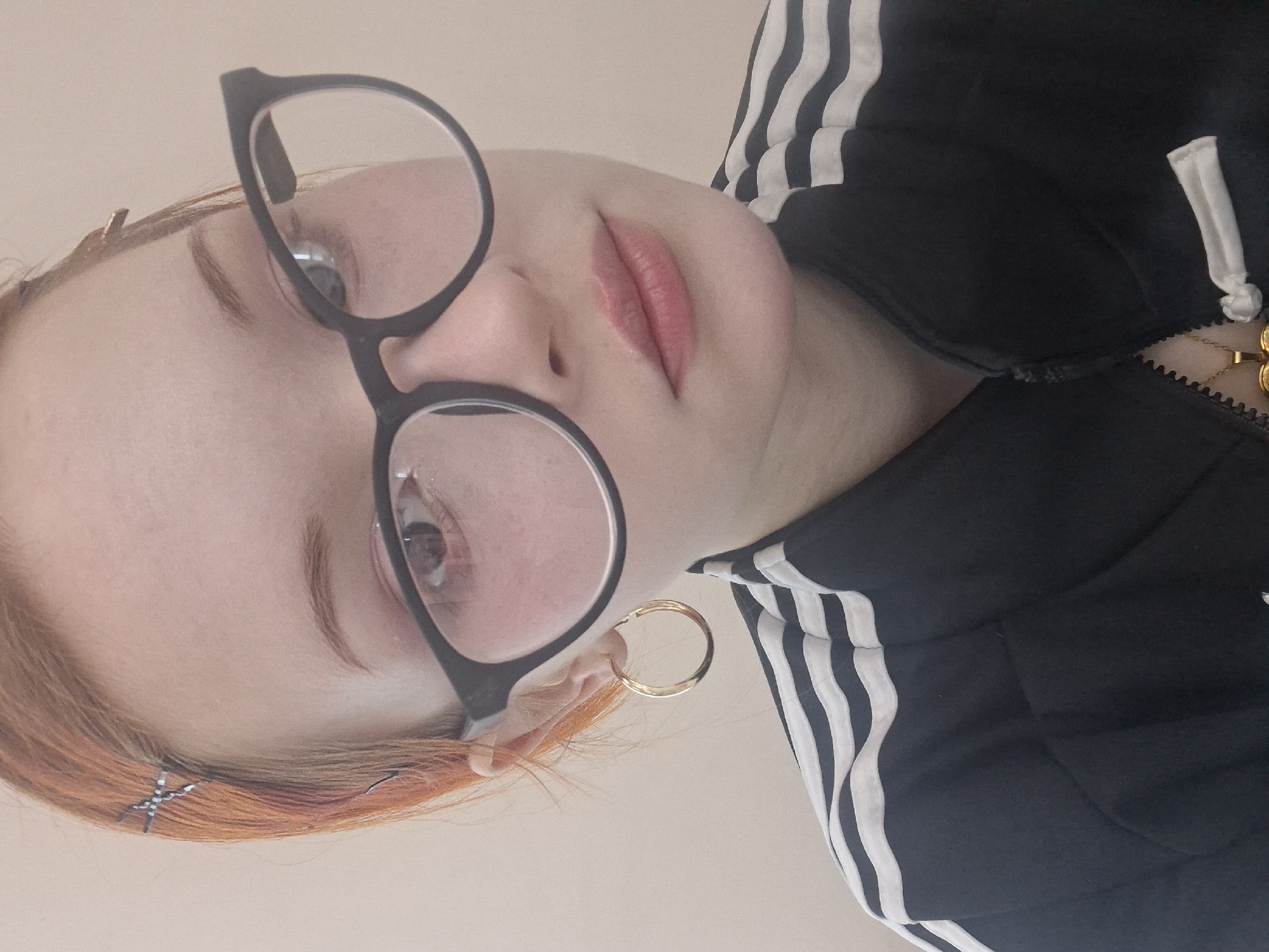 A person with reddish-orange hair is wearing black framed glasses and a black Adidas jacket with white stripes on the sleeves. They have a gold hoop earring in their left ear and a silver hairpin in their hair. They are wearing lipstick. The background is a solid, light beige color. The image appears to be a close-up selfie taken indoors. There is no information about the location being Chaikovsky, Russia within the image.