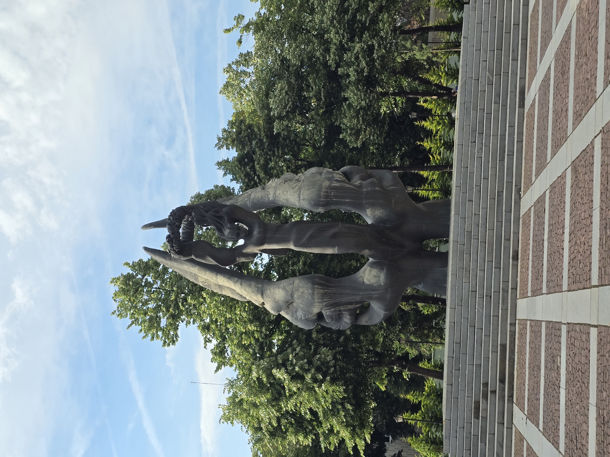 The image captures a large, dark-colored abstract sculpture set within an outdoor public space. The sculpture, which occupies the central and left portion of the frame, features flowing, intertwined forms that suggest a dynamic, upward movement. These forms appear organic and somewhat resemble hands or elongated, stylized human figures reaching or converging, with intricate details visible at the uppermost section, possibly forming a crown-like element or entangled shapes.
Behind and around the sculpture, lush green trees with dense foliage create a natural backdrop. Above the trees and the sculpture, a bright blue sky with scattered wispy white clouds is visible, indicating clear and sunny weather, likely during the day. To the right of the sculpture, a paved area is visible. This area includes a section of grey, stone-like steps or risers running vertically (when correctly oriented) along what appears to be a wall or raised platform. Adjacent to this, there is a patterned ground surface made of reddish-brown, possibly brick or cobblestone, material interspersed with lighter, vertical bands, forming a grid-like design. No human activity or visible text is present in the image.