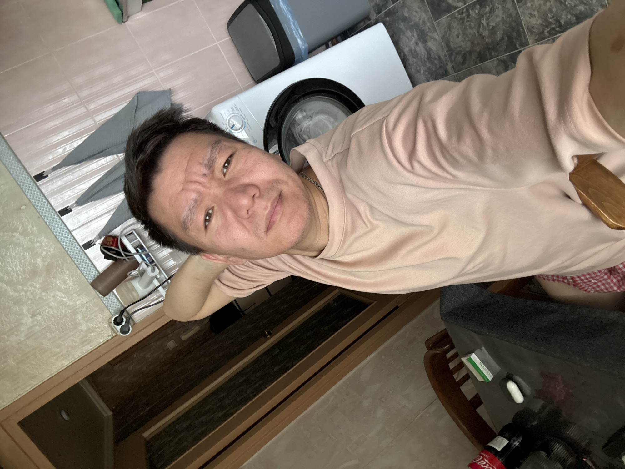 The image shows a man taking a selfie in a room. He is wearing a light pink t-shirt and has short, dark hair. His expression is a slight grimace. In the background, a white washing machine is visible, along with a grey trash can. The room appears to be a bathroom or laundry area, with tiled flooring and a wooden cabinet. There are also some electrical outlets and cords visible, suggesting the presence of appliances. A blanket with a red and white checkered pattern is draped over a piece of furniture. On a dark surface in the foreground, there are a few small items, including a green box with some white text and a white capsule-shaped object. A portion of a Coca-Cola bottle is also visible. The lighting suggests it is daytime. The scene is likely indoors in a residential setting in Khimki, Russia.