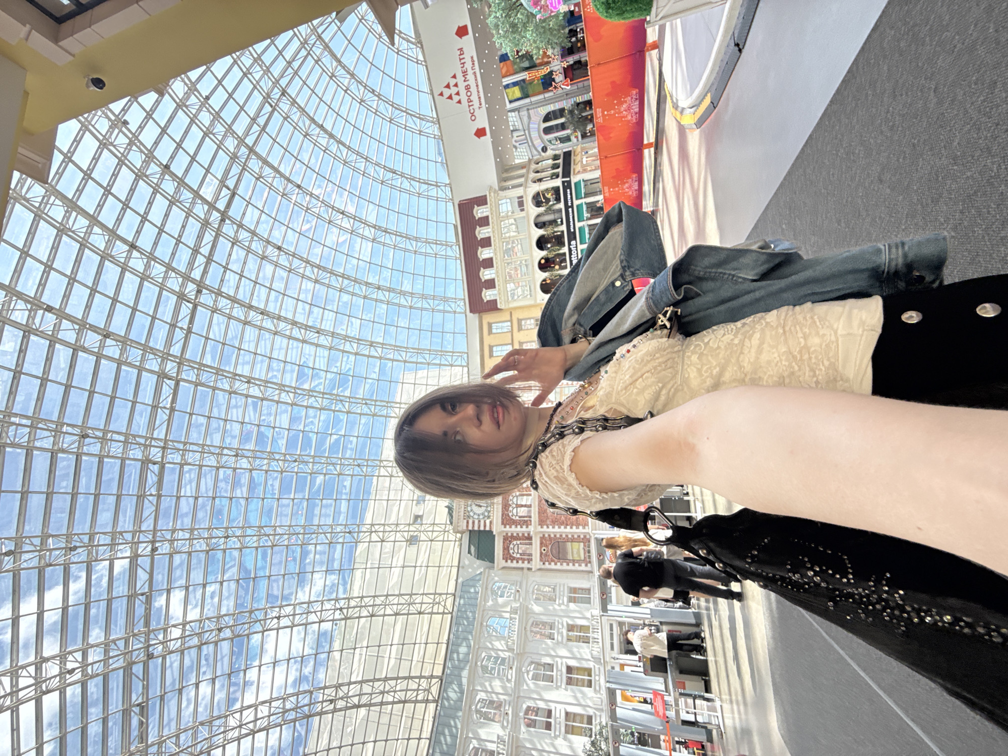 The image is taken from a low angle inside a large indoor space featuring a glass dome ceiling. The person taking the photo is holding the camera up and to their side, showing their arm and body in the foreground. They are wearing a light-colored, lacy top and a dark garment with studded details. A denim jacket is draped over their clothing.
In the background, a simulated European city streetscape is visible, complete with building facades and signage in Russian script, including "ОСТРОВ МЕЧТЫ" (Island of Dreams) indicating that the location is likely the "Dream Island" theme park in Moscow, Russia. People are walking on the pathway between the camera and the simulated street scene. The flooring is a neutral gray. The distinctive architecture of the glass dome is visible, composed of numerous intersecting metal supports forming a grid pattern.