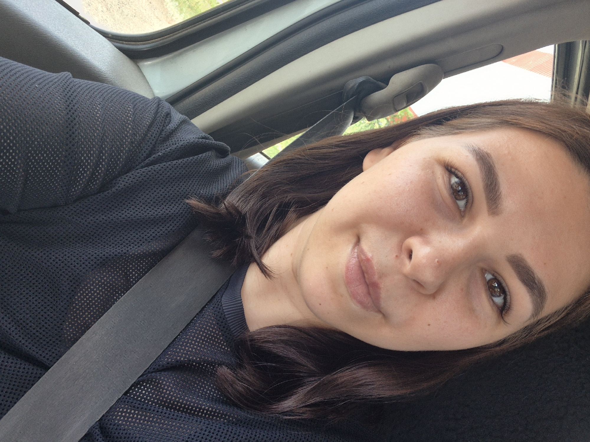 A young woman with dark brown hair, styled in loose waves, is captured in a close-up, frontal selfie taken from inside a vehicle. She is wearing a dark, sheer, long-sleeved top over a black undershirt. A grey seatbelt is prominently fastened across her chest. The background shows the interior of a car, with a window on the left revealing a blurred outdoor scene. The lighting suggests it is daytime. There is no discernible location information for Chaikovsky, Russia.