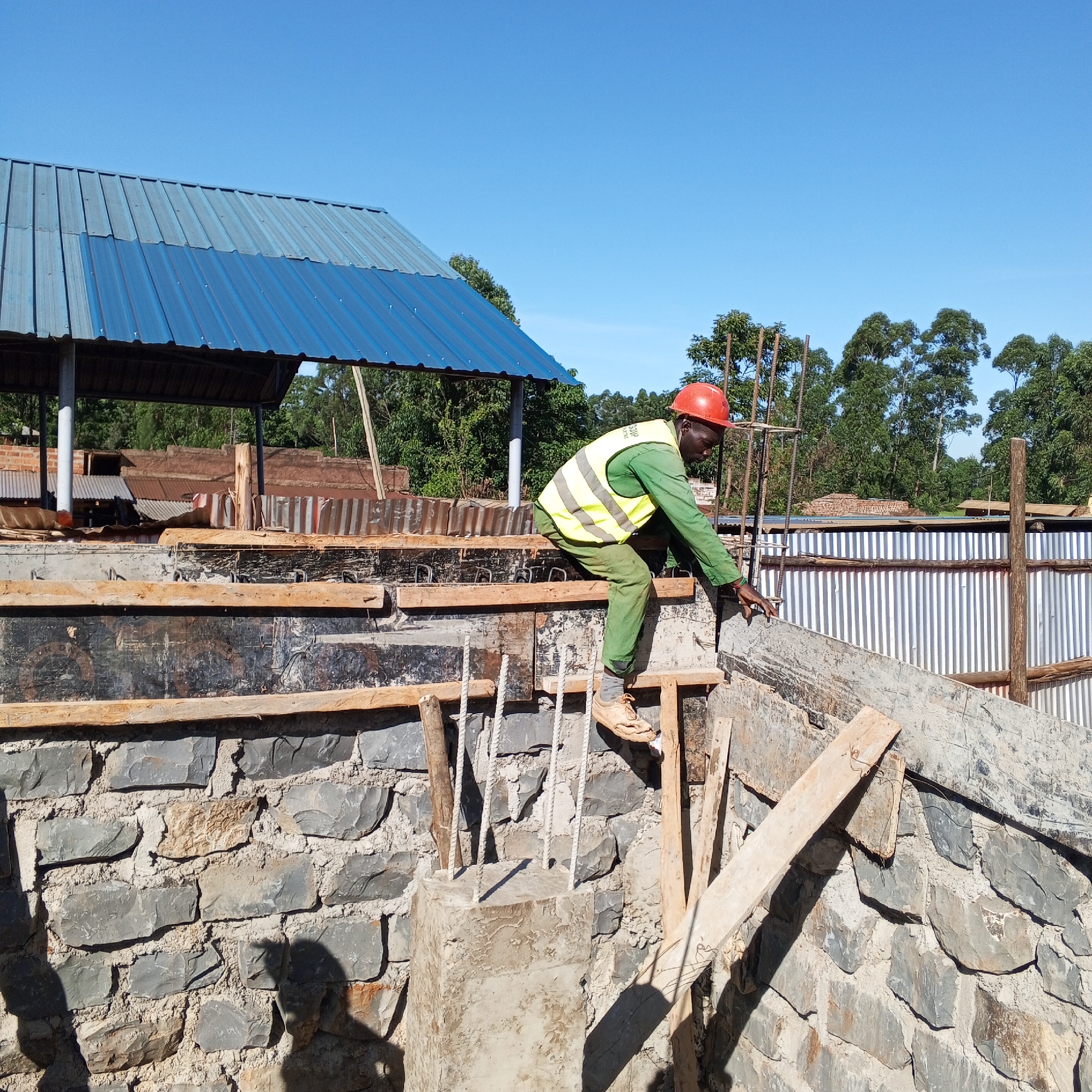 In Borabu Chitago ward, Kenya, a construction worker wearing a red hard hat, a fluorescent yellow safety vest over green clothing, and tan shoes is positioned atop a partially constructed stone wall. He appears to be inspecting or adjusting the framework. The wall consists of irregularly shaped grey stones. Wooden planks and concrete forms are used to shape the structure, with reinforcing steel bars visible. In the background, a blue and grey corrugated metal roof covers an open structure. Beyond that, trees and other buildings are seen under a clear blue sky.