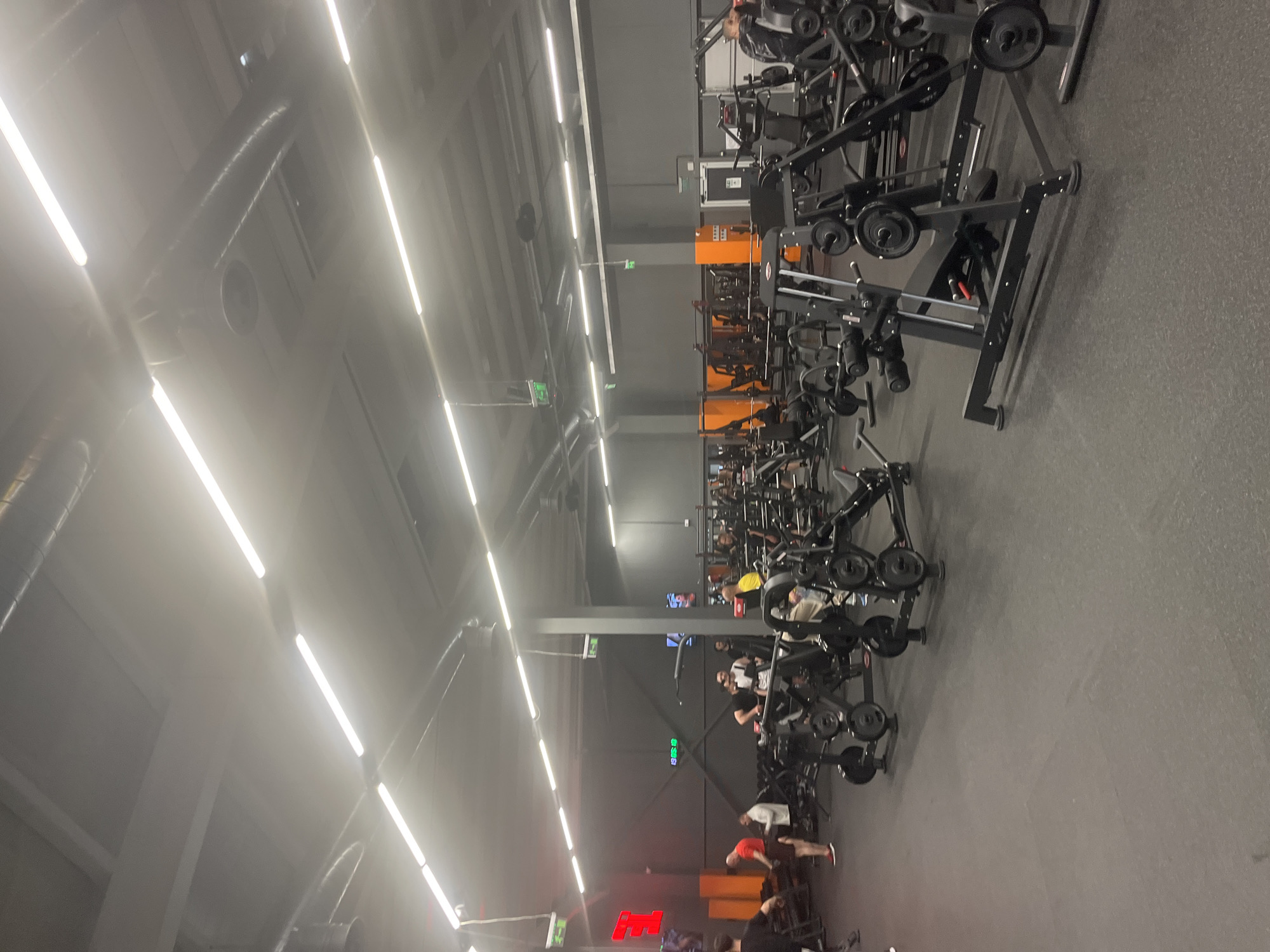 The image shows the interior of a gym. There are rows of weight machines extending into the distance. People are using the equipment. The gym appears to be in Plovdiv, Bulgaria, based on visual identification of the "E" logo. The scene has artificial lighting. The floor is gray.