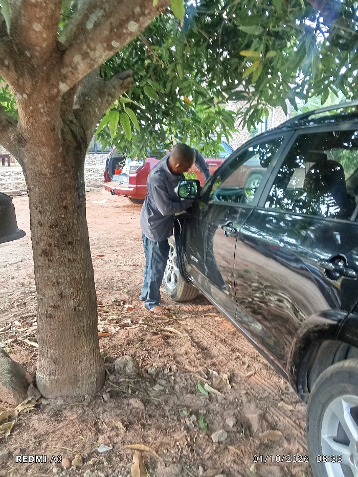 A man standing next to a black SUV appears to be adjusting the side mirror, captured outdoors amidst trees and foliage. The environment seems to be a dirt yard or rural setting, with another red vehicle partially visible in the background. A large tree trunk is positioned to the left, close to the SUV. The timestamp indicates a date in 2026.