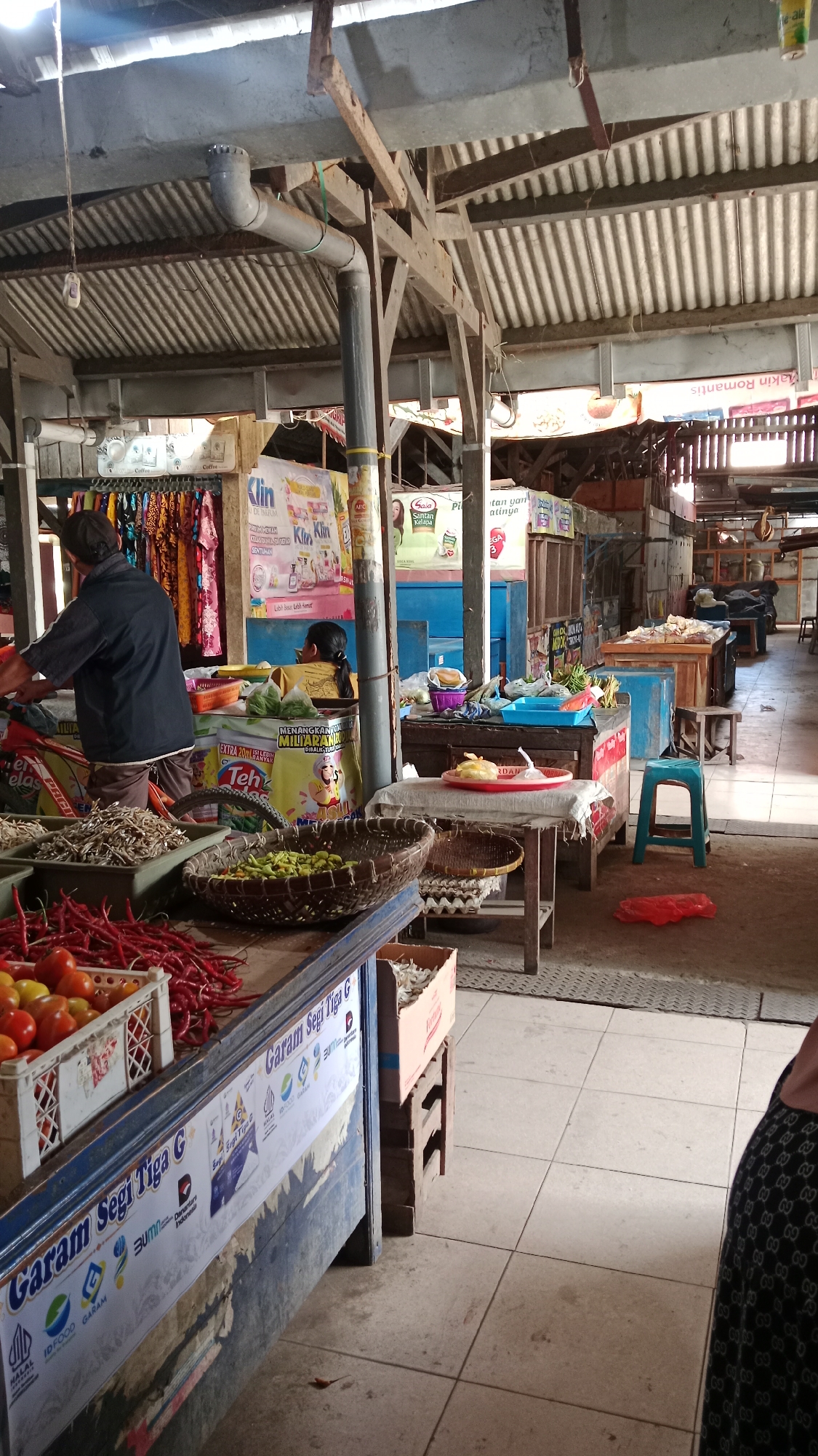 The image shows an indoor market scene in Kademangan, Indonesia. In the foreground, a stall displays fresh produce, including red chili peppers in crates and a basket of green chili peppers. Behind the produce, a man wearing a dark shirt and cap is actively working, and a woman with her back to the camera is also present at the stall. Further into the market, various goods are arranged on tables and shelves, including clothing and packaged items. The market appears to have a corrugated metal roof supported by wooden beams, and the floor is tiled. The overall impression is of a bustling local marketplace.