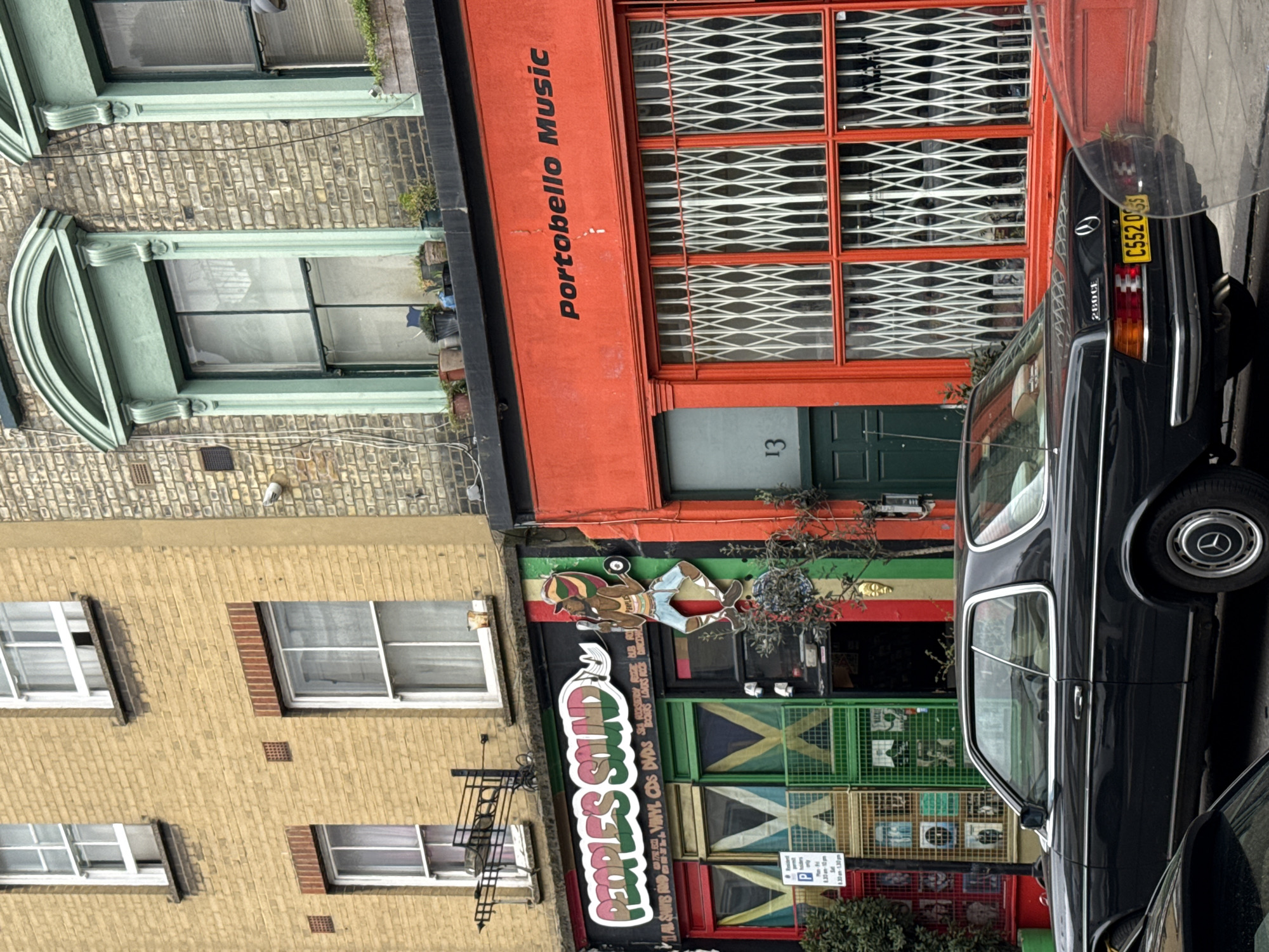 The image depicts a street scene on Portobello Road in London, United Kingdom. Two multi-story brick buildings house ground-floor retail establishments.
The building on the left, constructed of light yellow brick, features four white-framed sash windows with dark sills on its upper floors. Its ground floor hosts "People's Sound," a shop with a facade patterned in vertical stripes of red, green, black, and yellow. The shop's name is rendered in large white letters outlined in green and red. A stylized wooden cut-out of a man wearing a Rasta-style hat, holding a microphone and a record, is affixed to the right of the sign. The shop windows are protected by metal grates, some of which feature a yellow 'X' pattern on a green background, displaying vinyl records or album covers behind. A white parking sign detailing permit restrictions is visible on a pole to the left of the shop entrance.
The building on the right, constructed of darker brick, features two upper-floor windows with ornate arched, light green decorative surrounds. A black ledge above the ground-floor shop holds several potted plants. The ground floor is occupied by "Portobello Music," identifiable by its vibrant orange facade with the shop's name printed in black capital letters above the main windows. These windows are secured by white collapsible security grilles. A dark green door, marked with the number "13" in white, is situated to the left of the grilles.
A black Mercedes-Benz 280CE coupe, bearing the UK license plate C552 OSS, is parked on the street in the foreground, partially obscuring the lower sections of the storefronts. Its light-colored interior is visible. A portion of a second dark vehicle is visible in the extreme left foreground. Overhead utility wires are visible against the brick facade of the right building, and a golden decorative mask is mounted on the wall between the two ground-floor businesses, above the parked car.