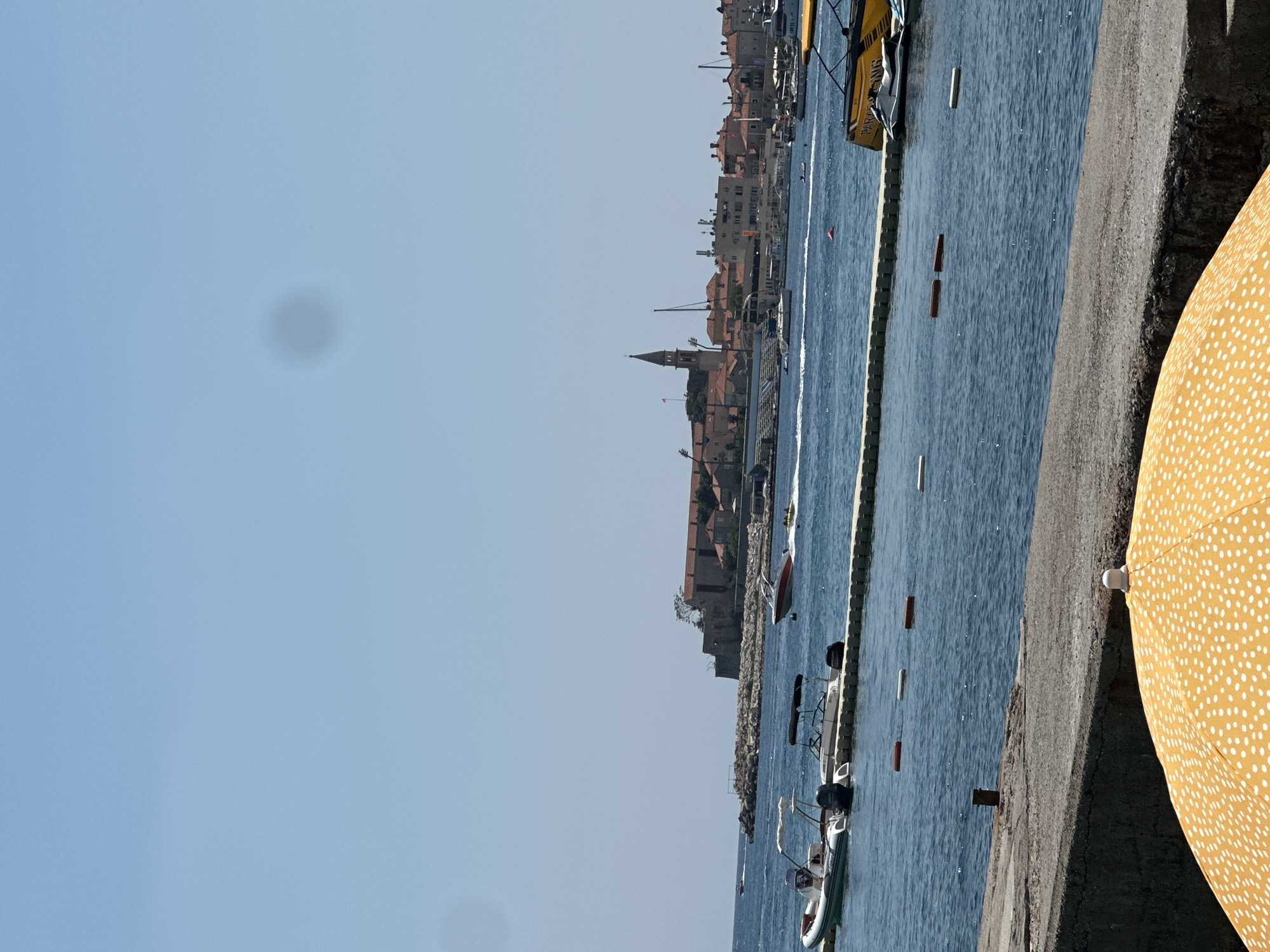 The image depicts a sunny day in Budva, Montenegro, with a clear blue sky overhead and calm blue waters of the Adriatic Sea. In the foreground, the top portion of a bright yellow umbrella with white polka dots is visible, suggesting a beach or waterfront setting. A concrete barrier runs along the edge of the foreground, separating it from the water.
The middle ground features several boats docked along a floating walkway, including inflatable boats and speedboats. Red and white buoys are scattered in the water, marking a swimming or boating area. Beyond the boats, a town with red-tiled roofs and a prominent church steeple is visible, situated on a peninsula. The historic architecture suggests the old town of Budva.
In the background, more boats are visible on the water, and some people are engaged in water sports, such as tubing behind a speed boat. The overall scene is one of leisure and activity in a picturesque coastal city.
