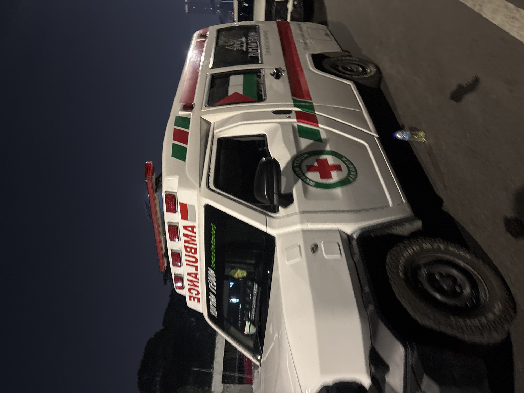 An overturned, white ambulance with red and green stripes is parked on a street at night. The word "AMBULANCE" is written in reverse on the side of the vehicle. A Palestinian flag is visible through the rear window. A red cross symbol with the word "JAKARTA" is on the side of the ambulance. A bottle of water sits on the ground next to the vehicle. The scene appears to be in Central Jakarta, Indonesia.