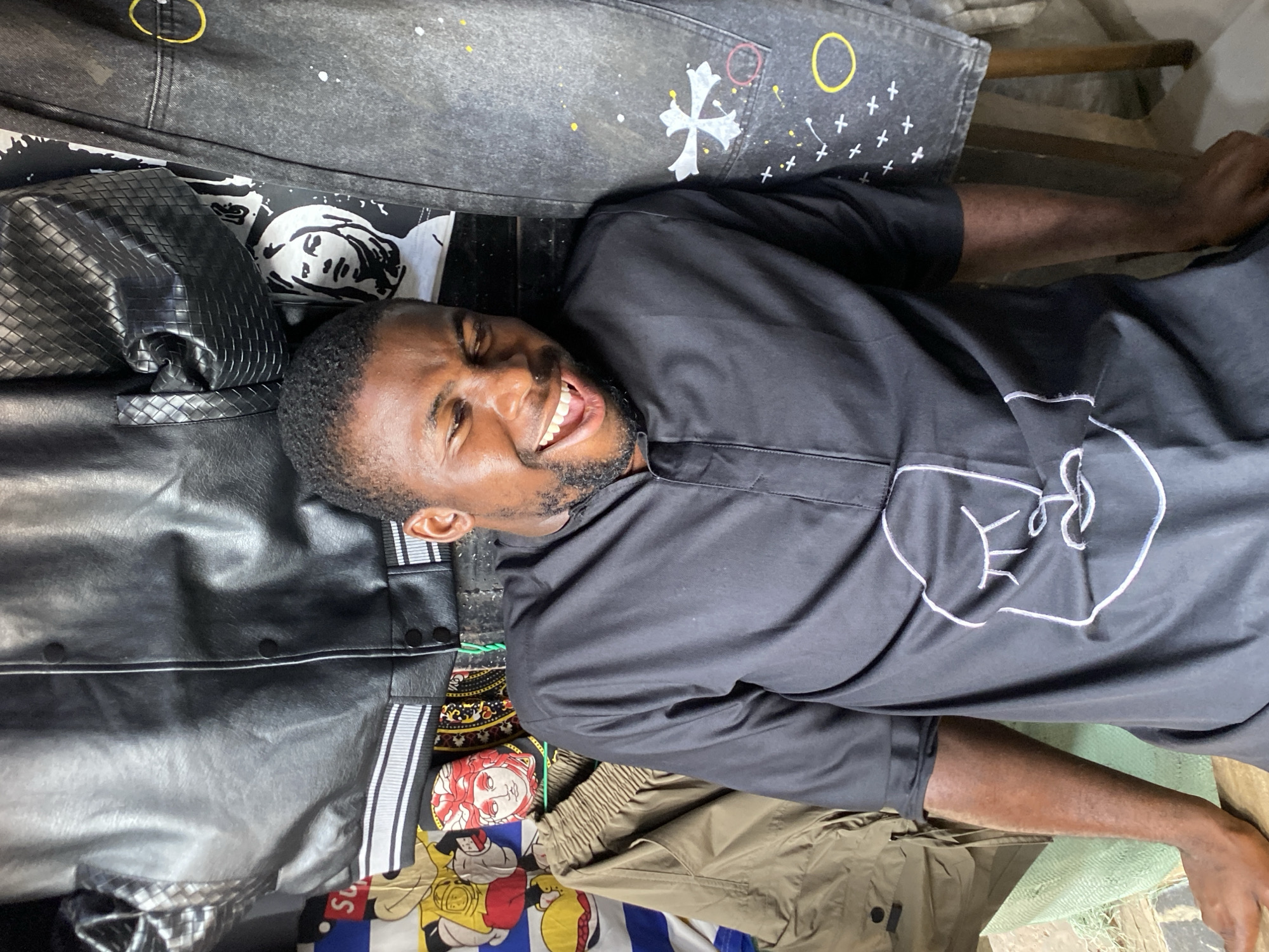The image captures a smiling man, likely of African descent, lying amidst a vibrant collection of clothing and accessories. He has short dark hair and a beard, and his mouth is wide open in an expression of laughter or happiness as he looks towards the camera. He is wearing a black short-sleeved shirt that features a distinctive white abstract design, possibly a stylized face or fist, on the chest.
Around him, a variety of items are arranged, creating a cluttered yet intriguing scene. To his left, near his head, is a black quilted bag or satchel, alongside a black and white patterned fabric displaying abstract faces or figures. Above him, a pair of dark grey or black denim jeans is visible, adorned with white crosses, white paint splatters, and small yellow circles and crosses.
Below and to his right, other garments include a pair of brown or khaki pants, and a black leather-like jacket with white stripes running down the side. A colorful fabric with cartoon characters, including what appears to be Mickey Mouse and a partial Superman logo, is also visible. A light green woven mat or bag is partially visible under his right arm, and wooden structures can be seen in the background, hinting at a rustic or makeshift setting.
The scene appears to be indoors, possibly a market stall, a shop, or a personal space filled with merchandise. The man's cheerful demeanor is a central element, conveying a relaxed and jovial mood within this environment in Bukuru, Nigeria.