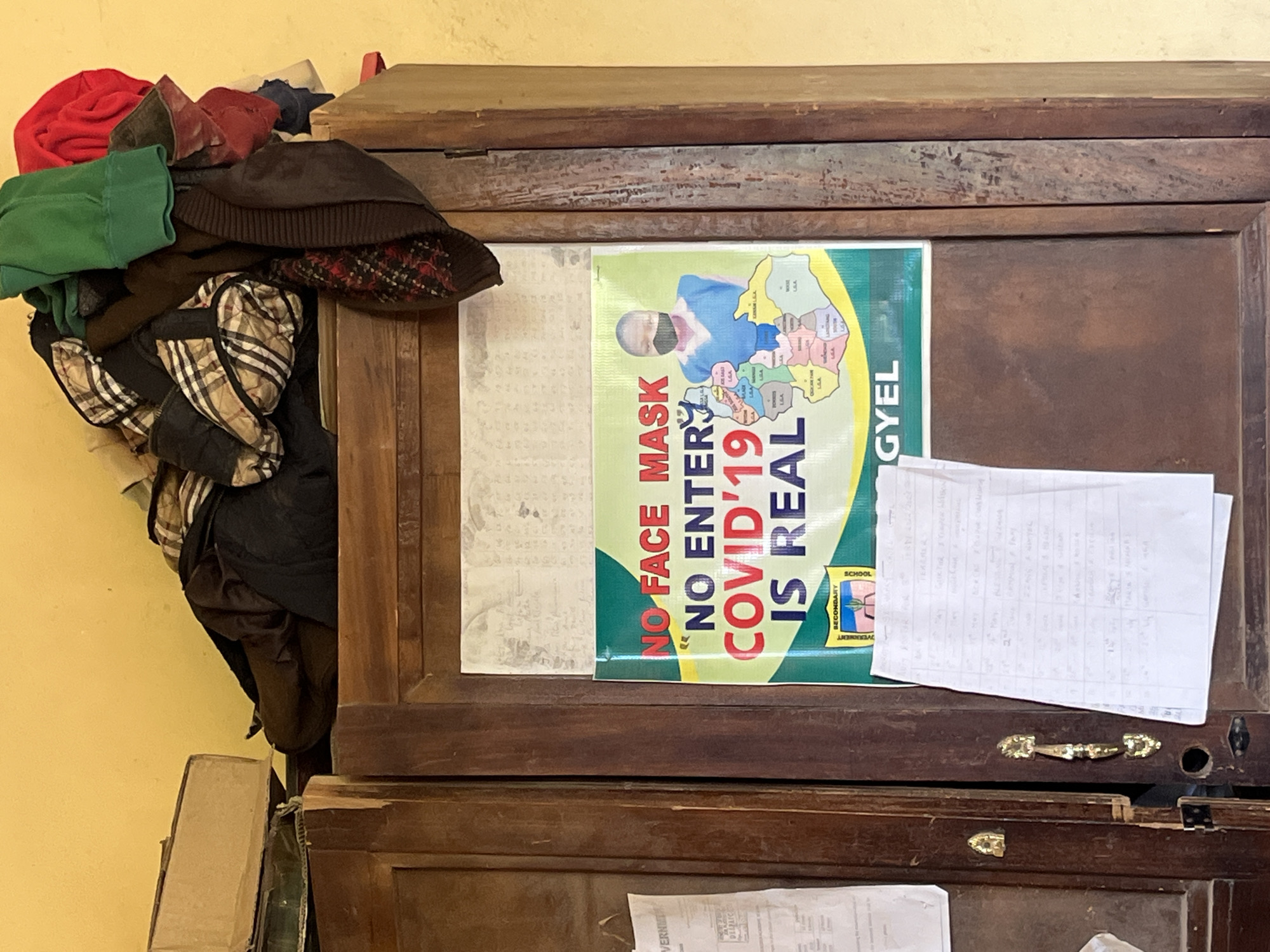 This image depicts the exterior of a wooden cabinet, likely in a public space within Bukuru, Nigeria. Piled atop the cabinet are various items of clothing, including a plaid jacket, a red garment, and a green hooded top. A prominent sign is affixed to the cabinet door, featuring a graphic of a person wearing a face mask and a map of local government areas. The sign's text reads "NO FACE MASK NO ENTER COVID'19 IS REAL". Below this sign, a piece of paper with handwritten notes is attached to the cabinet. The cabinet itself shows signs of wear and tear, with visible damage to its surface and hardware. The surrounding wall is painted a muted yellow.