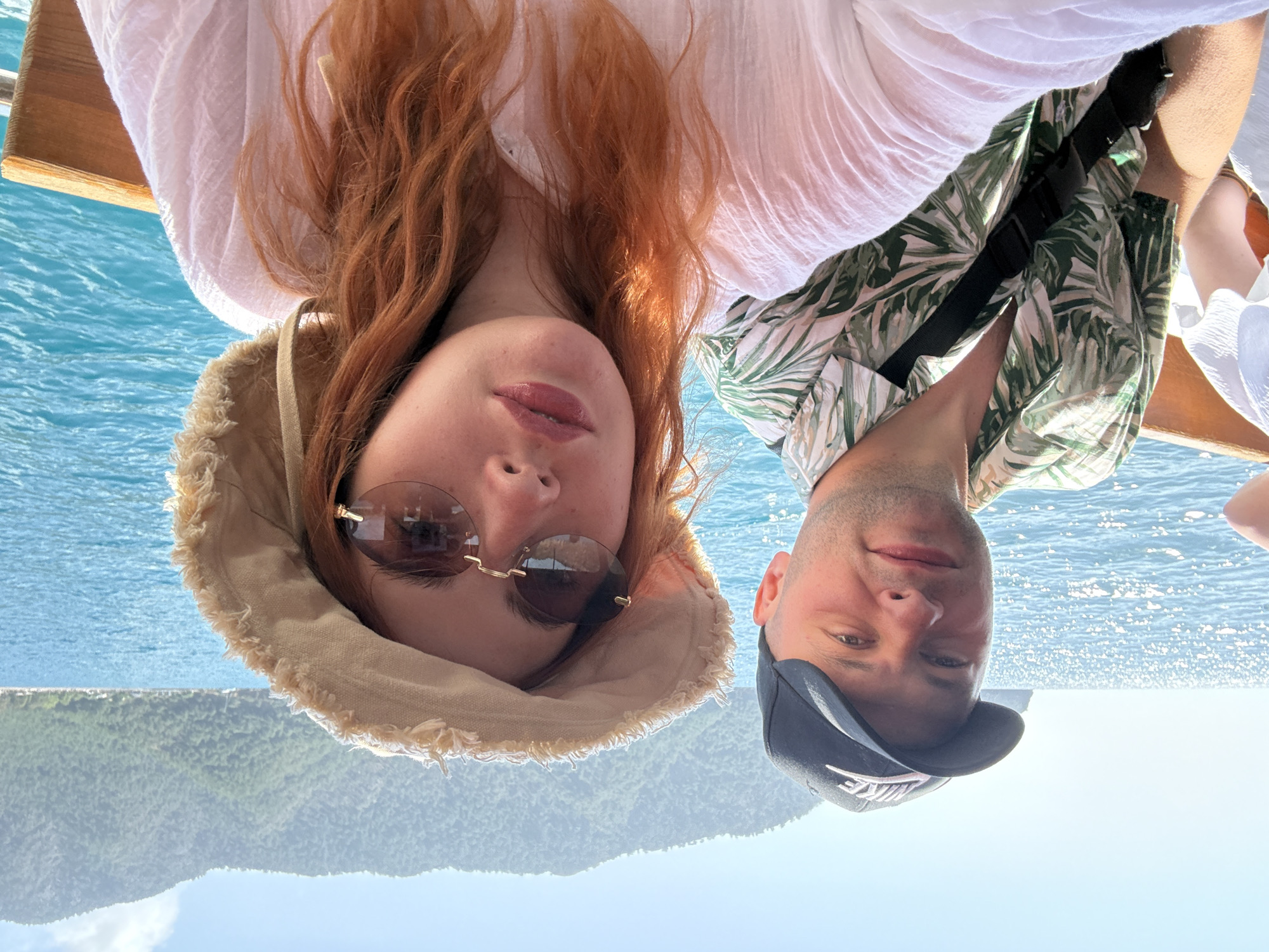 Two individuals are positioned closely, facing the camera in an upside-down orientation. The environment features blue water and a hazy, mountainous backdrop, suggestive of an outdoor setting, possibly a boat excursion.
The person on the left has red hair, wears a neutral-colored sun hat and rimless sunglasses, and is dressed in a white top. The person on the right wears a dark baseball cap and a light-colored, tropical-patterned shirt.
The geographical location, Kumluca, Turkey is inferred from available information about the imagery, it is unclear what is their action or interaction in the image.