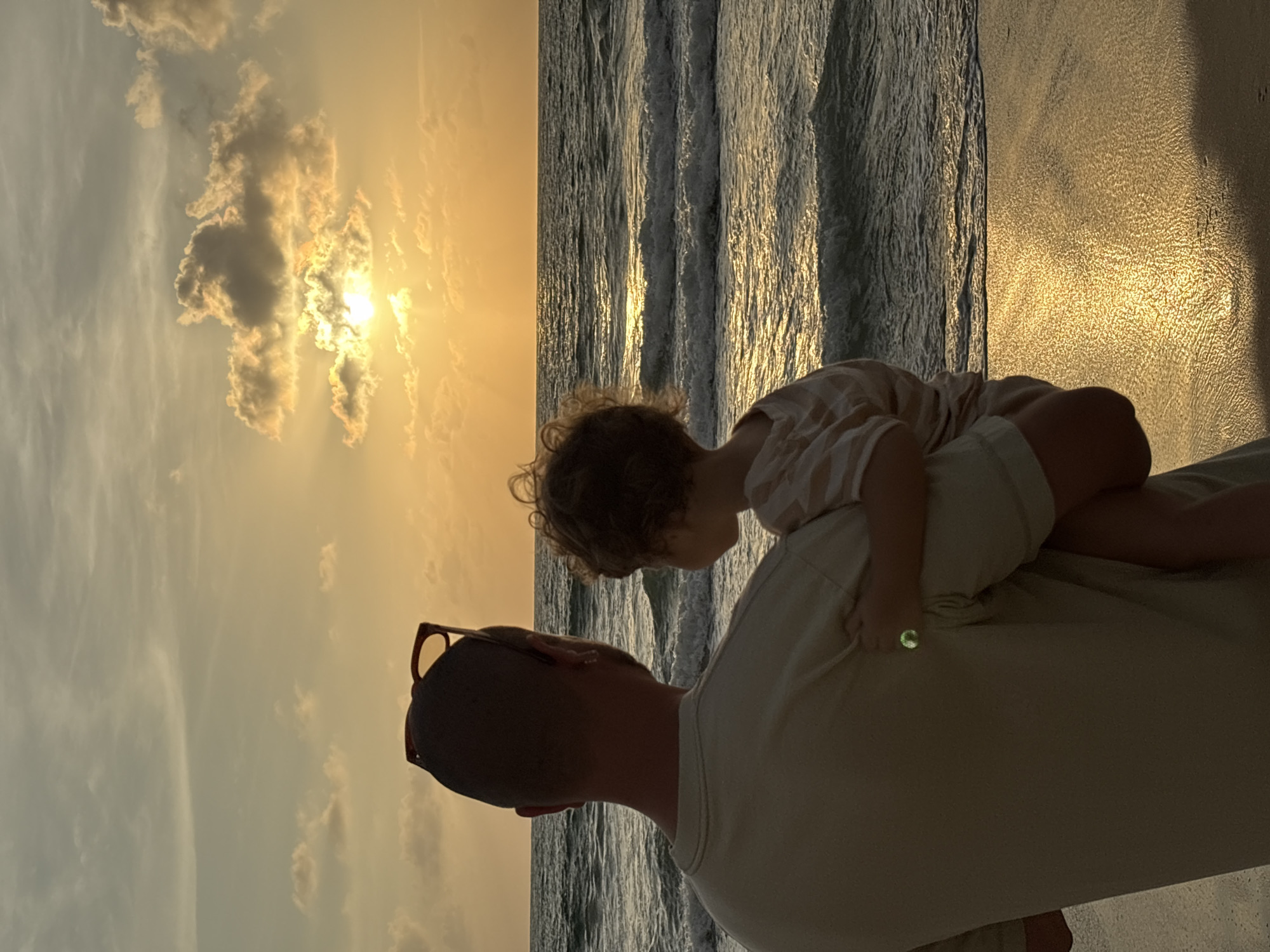 A man stands on a beach in Choeng Thale, Thailand, holding a young child in his arms, both facing the ocean. The setting sun casts a warm, golden glow across the sky and reflects on the water's surface, with gentle waves lapping at the shore. The man has sunglasses perched on the back of his head, and the child is wearing a striped shirt. The overall atmosphere is one of quiet observation and shared moment between the two individuals against the backdrop of a serene beach sunset.