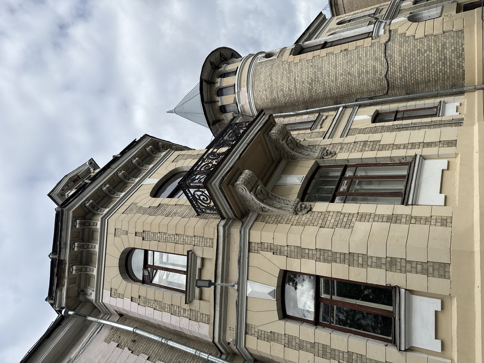 The image depicts an exterior view of a building with a distinctive architectural style. The building is characterized by its beige facade, accented with lighter beige stone blocks and yellow brickwork. A prominent feature is a circular tower with a conical gray roof, resembling a turret. Several windows are visible, some with arched tops and dark wooden frames. A small balcony with ornate black metal railings is attached to the main building. The roofline features decorative cornices and eaves. A metal downspout runs vertically along the side of the building. The sky above is overcast with gray clouds. The scene is devoid of any people or visible activity, presenting a static architectural study. The building's architectural style suggests it is located in an older part of a European city, possibly dating back to the late 19th or early 20th century. Without additional information or visual cues, pinpointing the exact location within Moscow, Russia is not possible.