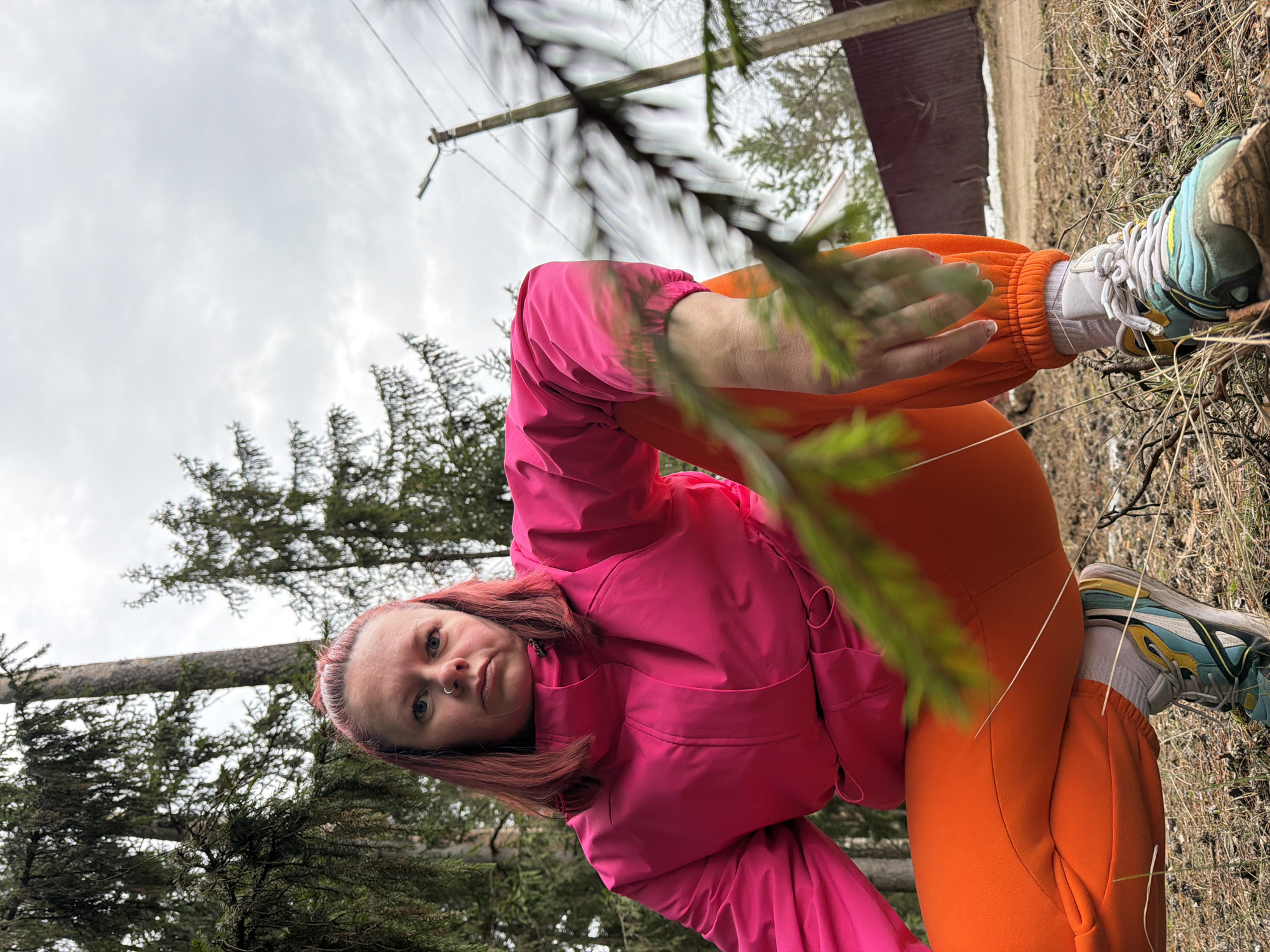 A woman is pictured outdoors in a wooded area. She wears a bright pink jacket and orange sweatpants. She is holding a small pine branch. Her shoes are white socks with blue, yellow, and white running shoes. The ground is covered in dry grass, twigs, and other forest debris. The sky is overcast. There are power lines in the background. It appears to be Chaikovsky, Russia.