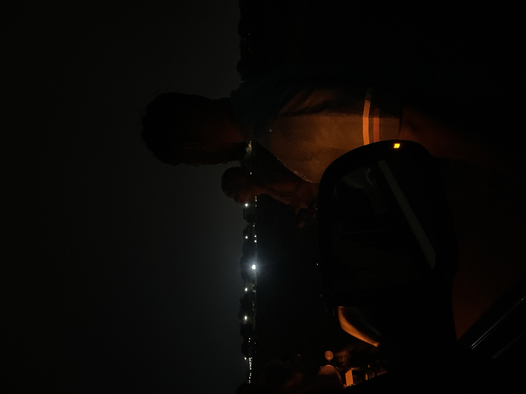 The image depicts a dimly lit scene, possibly at night. A person wearing a t-shirt with orange stripes is partially visible, facing downward. A chain with small lights hangs vertically, with a person looking down at it. A car's dashboard and part of the interior are in the foreground. Due to the limited visibility and lack of context, it is impossible to specify the exact location in Kuchingoro, Nigeria, or discern more detailed information about the actions or interactions.