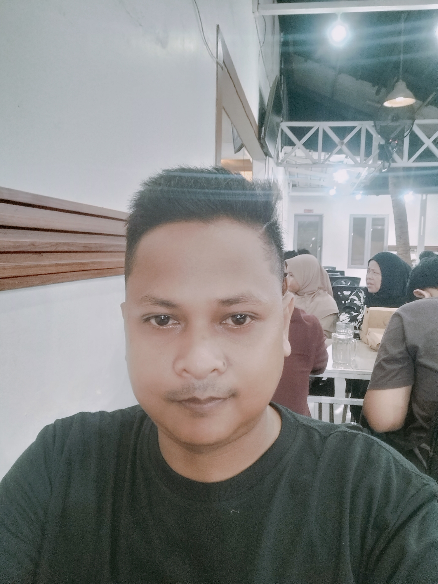 The image is a selfie taken by a man wearing a dark t-shirt in a restaurant or cafe. He is positioned in the foreground, with his face and upper body filling most of the frame. In the background, there are other patrons sitting at tables. One woman is wearing a light-colored hijab, and another woman is wearing a black hijab and a patterned top. There are tables, chairs, and glasses on the tables. The environment appears to be indoors with overhead lighting, suggesting it is either evening or the establishment has artificial lighting. The wall behind the man has horizontal wooden paneling. The ceiling structure above the patrons includes white cross beams and hanging lights, indicating a somewhat open or modern architectural design. The overall lighting is somewhat dim, with bright spots from the lights. The image quality is fair, with some blur and a slight yellow cast.