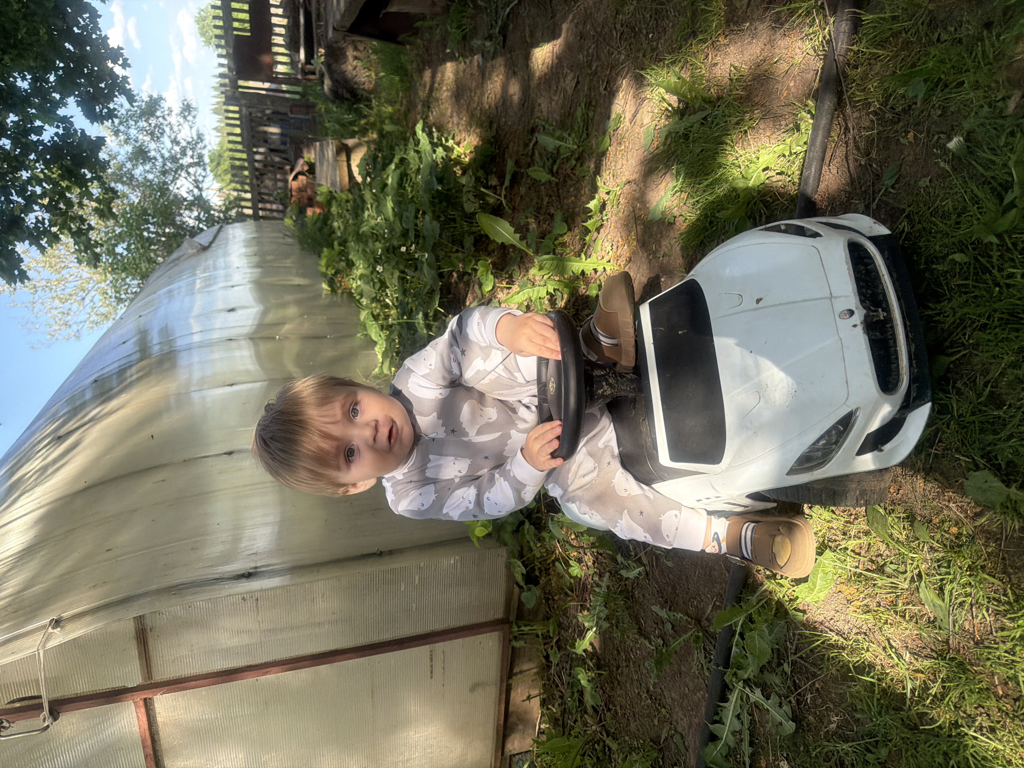 A young child, appearing to be between 1 and 3 years old, is sitting in a white toy car in a garden. The child is wearing a light-colored jumpsuit with a pattern of bears and stars, and brown shoes. The car is positioned on bare dirt and grassy patches, with green plants visible around the car. To the left, there is a greenhouse structure with translucent plastic panels. In the background, there's a wooden fence and more greenery. Given the request to incorporate the location of Panteleevka, Russia, it is presumed this is a backyard or garden within a residential area of that city. The scene appears to be taken on a sunny day, judging by the shadows and lighting. The child is looking at the camera with an open expression.