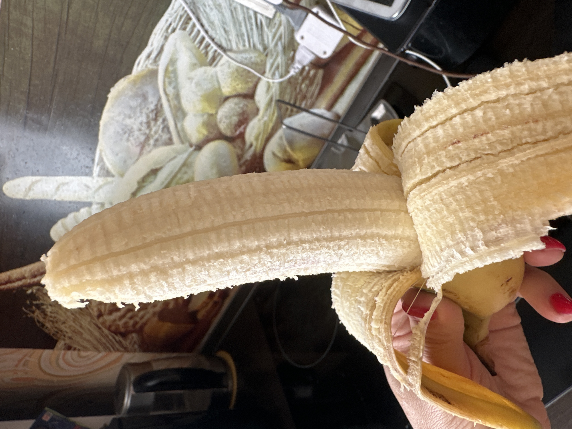A close-up shot shows a partially peeled banana held by a hand with red nail polish. The banana is light yellow and shows its textured flesh. In the background, a kitchen counter is visible with a black stovetop, a metallic object, and a wall with a decorative wallpaper depicting fruits and woven textures. Wires and a black appliance are also present. The image does not contain any information that would allow for the determination of its location in Novoaltaysk, Russia.
