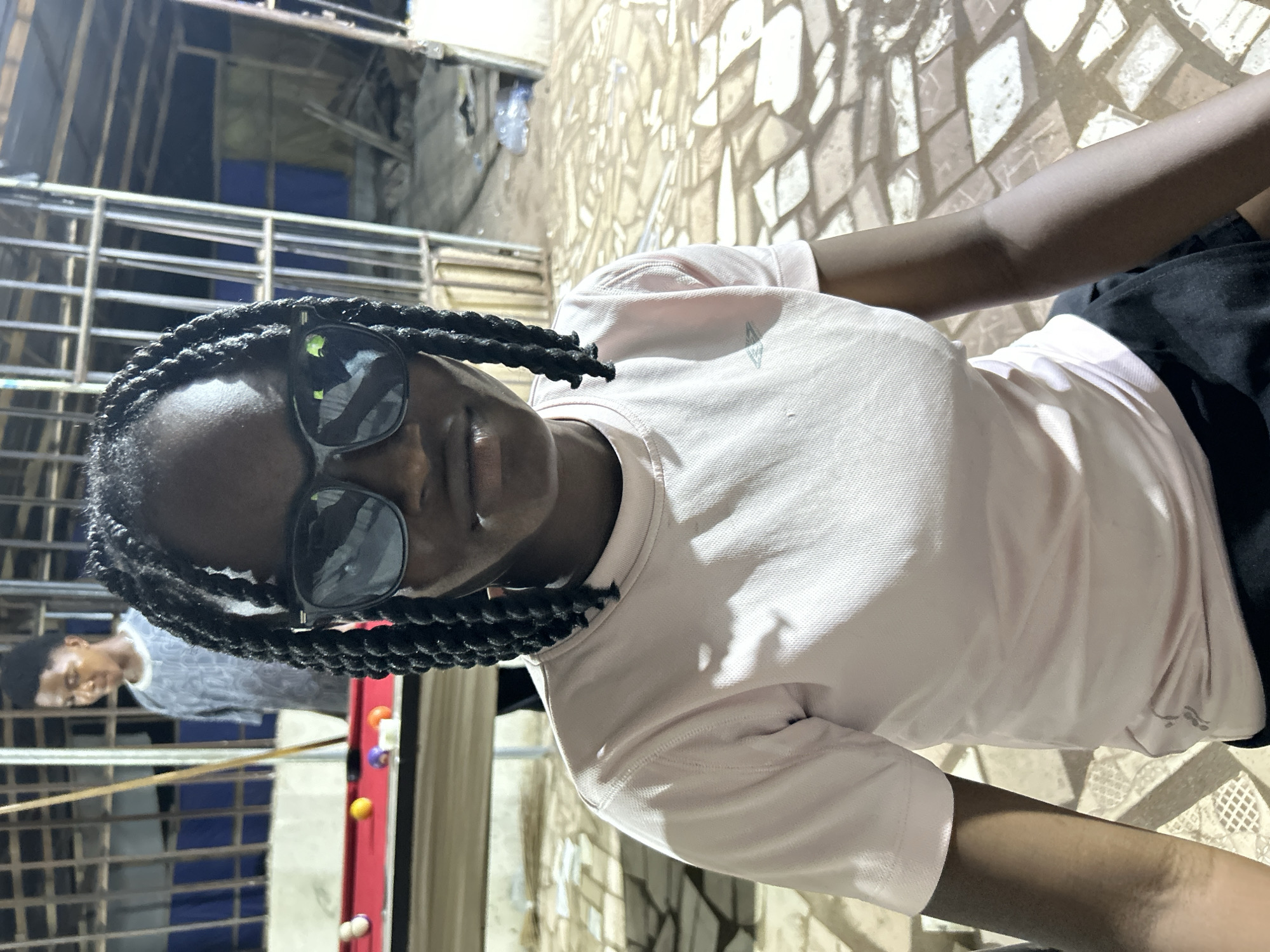 A person with dark skin, wearing dark sunglasses and a light pink shirt, is positioned close to the camera, their face angled slightly to the right. Their hair is styled in dark braids. Behind them, another individual is playing pool on a red pool table with colored balls. The pool table and player are partially obscured by a metal structure. The setting appears to be outdoors on a textured stone surface, under possibly dim lighting, possibly in the city of Ihiagwa, Nigeria.