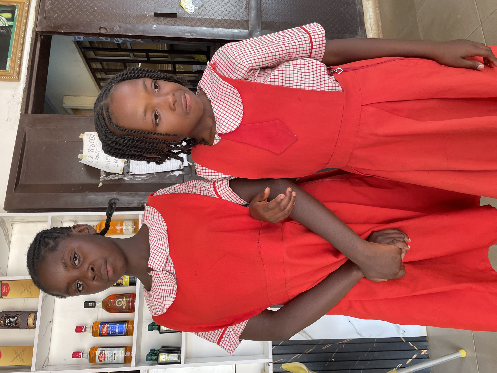 Two young girls, wearing matching red and white checkered school uniforms, stand in what appears to be an indoor setting, likely a shop or home. The girl in the foreground on the right has braided black hair and looks directly at the camera with a neutral expression, possibly a slight smile. Her red pinafore dress features a small red patch on the chest. The second girl, partially obscured behind the first, also has braided black hair and a similar neutral expression as she looks towards the viewer. She wears an identical uniform.
The background on the left features white shelving units displaying various bottled products, including several types of whiskey (e.g., "LUXOR WHISKEY," "ROYAL STANDARD WHISKEY") and rum (e.g., "CAPTAIN MORGAN RUM"), along with other unidentified spirits. On the right, a dark brown, possibly metallic, doorway or gate is visible, with a textured metallic surface above it. A gold-framed picture is partially visible on the far left.
A white piece of paper is taped to the dark doorway, displaying text that includes "32 88 032," "ONE POINT," "JOHN PE," and "GRA," possibly a phone number or identification details. The girls appear to be posing for a photograph. The lighting suggests it is daytime. The setting, attire, and subjects are consistent with the provided location context of Barakin Akawo, Nigeria. The floor consists of dark and light-colored tiles, and a yellow-tipped cane or pole can be seen on the ground in some cropped views.