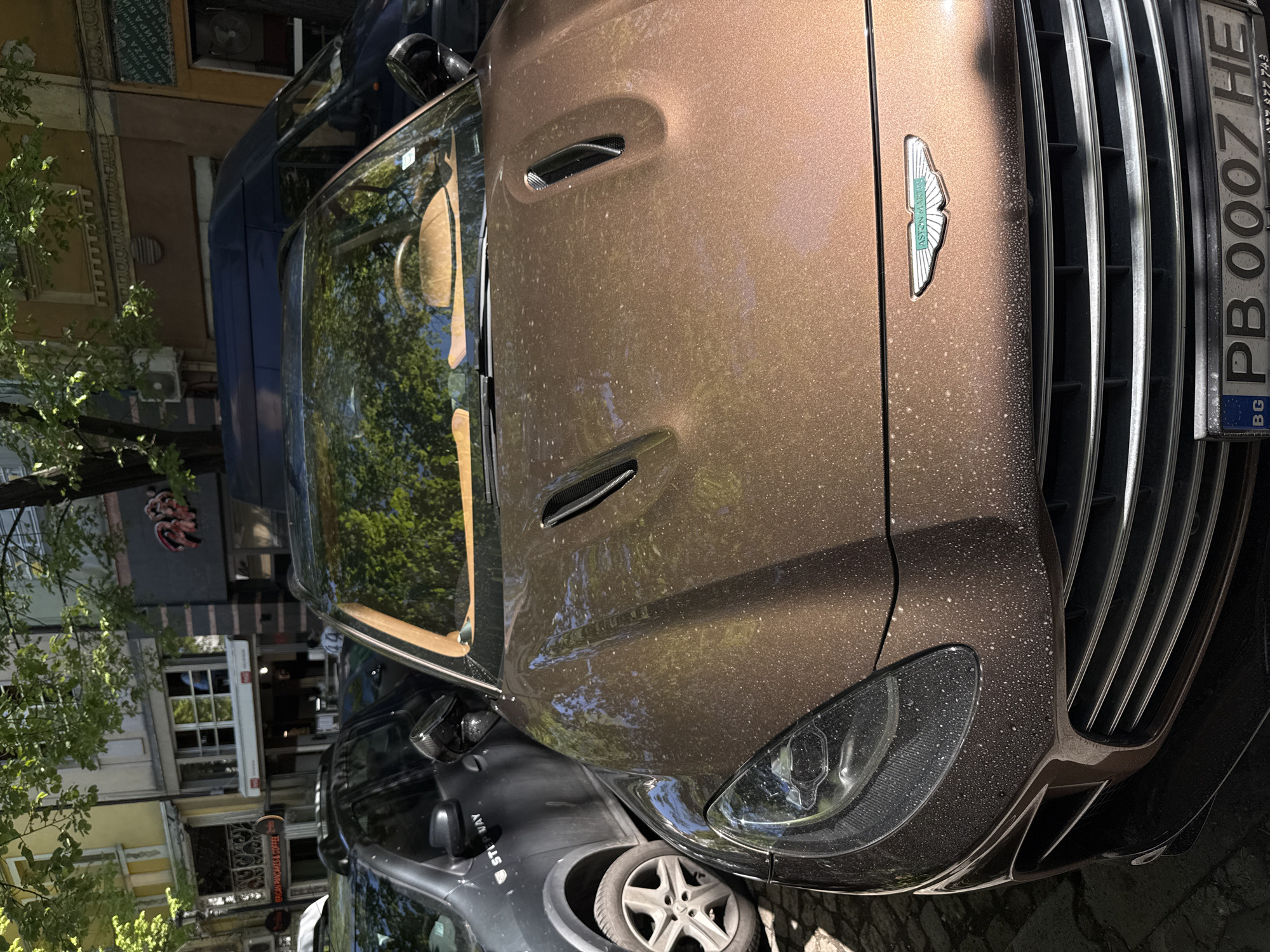 A close-up, canted-angle shot depicts the front end of a dirty, brown Aston Martin parked on a street. The car's hood, front grill, headlight, and license plate are visible. The license plate reads "PB 0007 HE" and identifies the car as being registered in Bulgaria (BG). Reflections in the car's windshield suggest a tree-lined street.
In the background, another parked grey vehicle is partially visible. A building facade, including windows and signage for "AFRICAN PANCAKES & COFFEE" is present. The sign "ПИЦА ПИЦА" (PIZZA PIZZA) can also be seen. Based on the signage, this scene is likely located in Plovdiv, Bulgaria.
