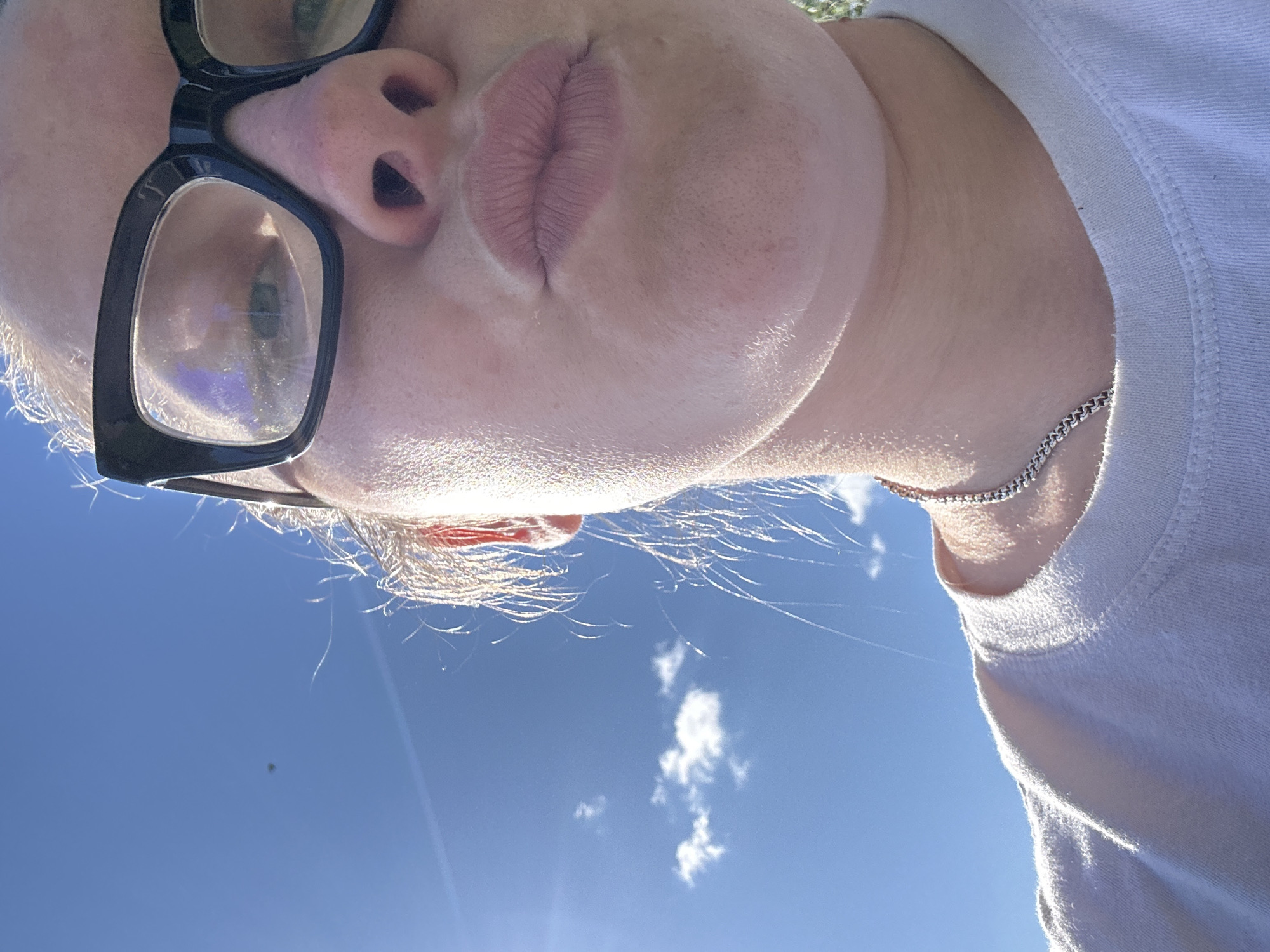 An extreme close-up selfie shows the upper part of a person's face and neck against a bright blue sky with a few white clouds. The person is wearing black-rimmed glasses and a white t-shirt. The sunlight is strong, creating lens flare and illuminating fine hairs around the person's face. The image is cropped tightly, focusing on the person's lips, nose, and glasses. There is no information to suggest this image was taken in the city of Калицино, Russia.