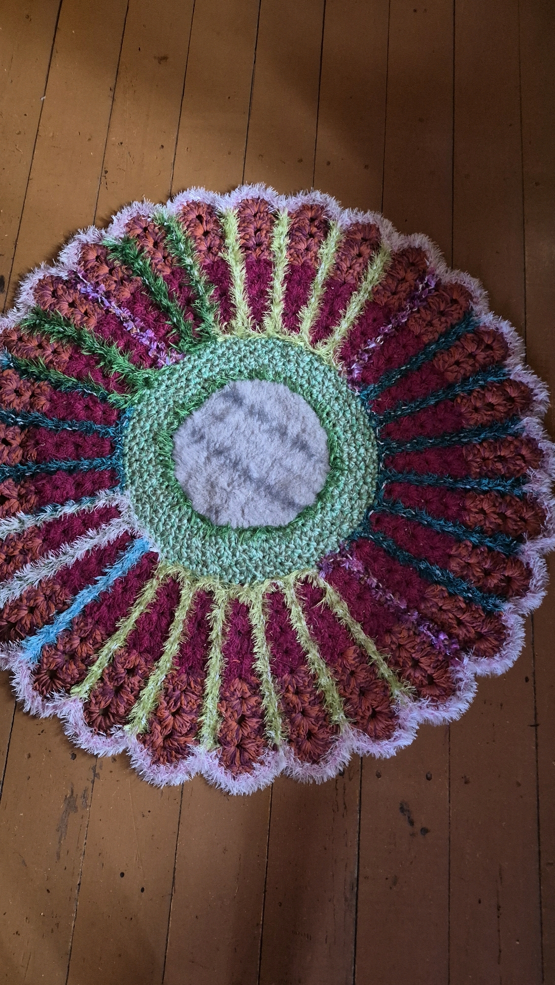 A circular, crocheted rug with a floral design lies on a wooden floor. The rug has a light gray fuzzy center, surrounded by a ring of green crochet. Radiating outward from the green ring are alternating petals of magenta and burnt orange yarn, each with a thin stripe of lime green yarn in the center. The outer edge of the rug is finished with a fluffy white and pink fringe. The wooden floorboards are worn and have a reddish-brown hue, with visible nails and scuffs. The lighting suggests an indoor environment. There are no people or animals present in the image. This scene could be located in a home in Ļūbaste 0001, Latvia.