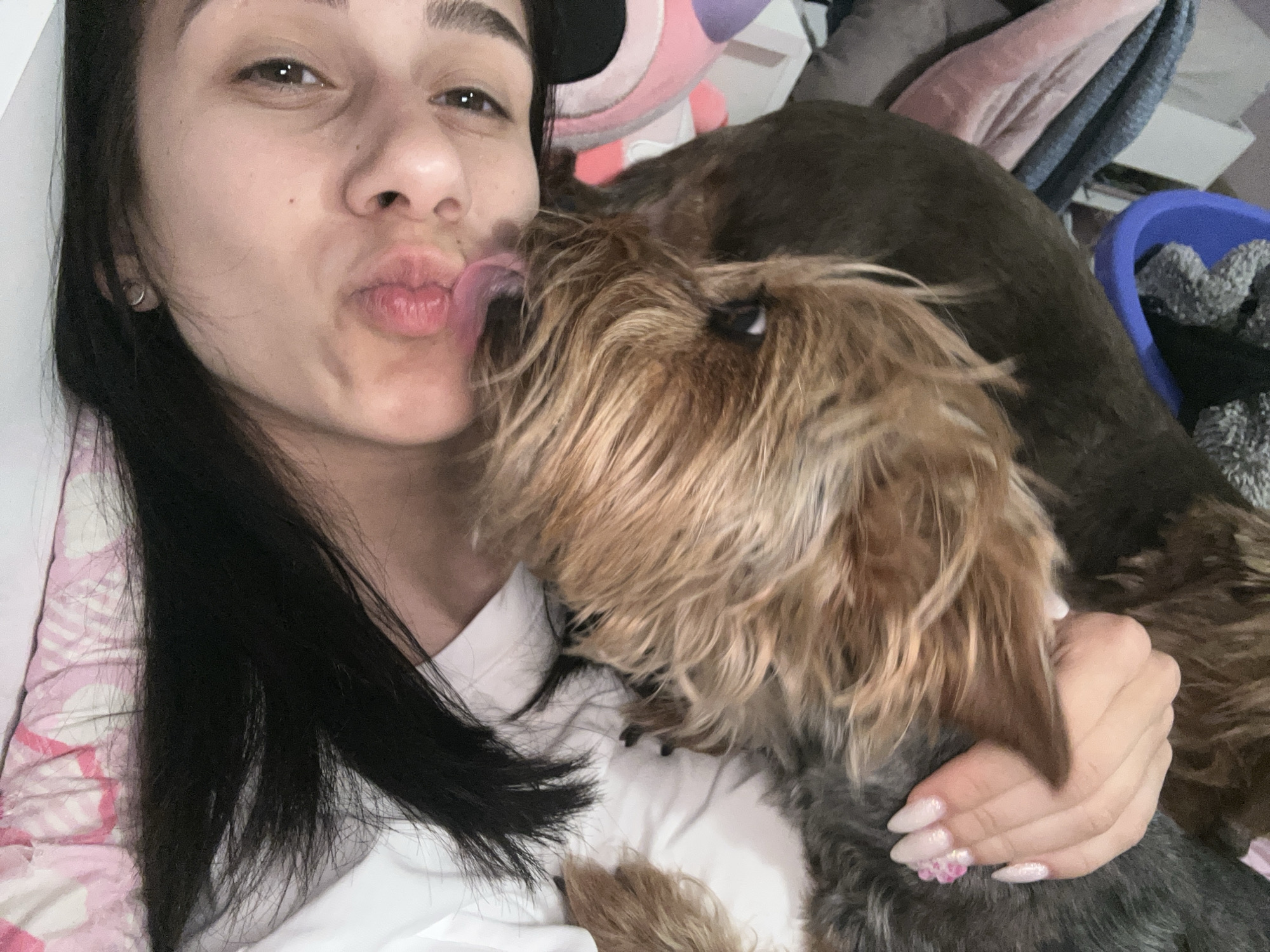 This close-up indoor shot captures a heartwarming moment between a young woman and her dog. The woman, with long dark hair and dark eyes, lies down, looking directly at the camera with a playful expression, puckering her lips as if for a kiss. Her right hand, adorned with light pink manicured nails featuring small flower designs, gently holds her pet.
Beside her, a small to medium-sized dog with shaggy, light brown, blonde, and darker grey fur is positioned closely. The dog is actively engaged, licking the woman's puckered lips or cheek with its pink tongue, conveying strong affection and playfulness. One of the dog's eyes is visible, looking towards the woman.
The setting appears to be a cozy indoor space, likely a bedroom or living area. A white fabric, possibly a shirt or bedsheet, covers the woman and supports the dog. To the left, a pink and white patterned fabric suggests bedding or a pillow. In the background, hints of other household items are visible: a dark brown blanket or large cushion, a partially obscured pink plush toy with an ear, and a blue plastic bin containing grey, folded items to the far right. The overall mood is intimate and affectionate, highlighting the special bond between the owner and her pet. There is no visible text in the image, and no specific time of day or location cues are apparent other than it being indoors.