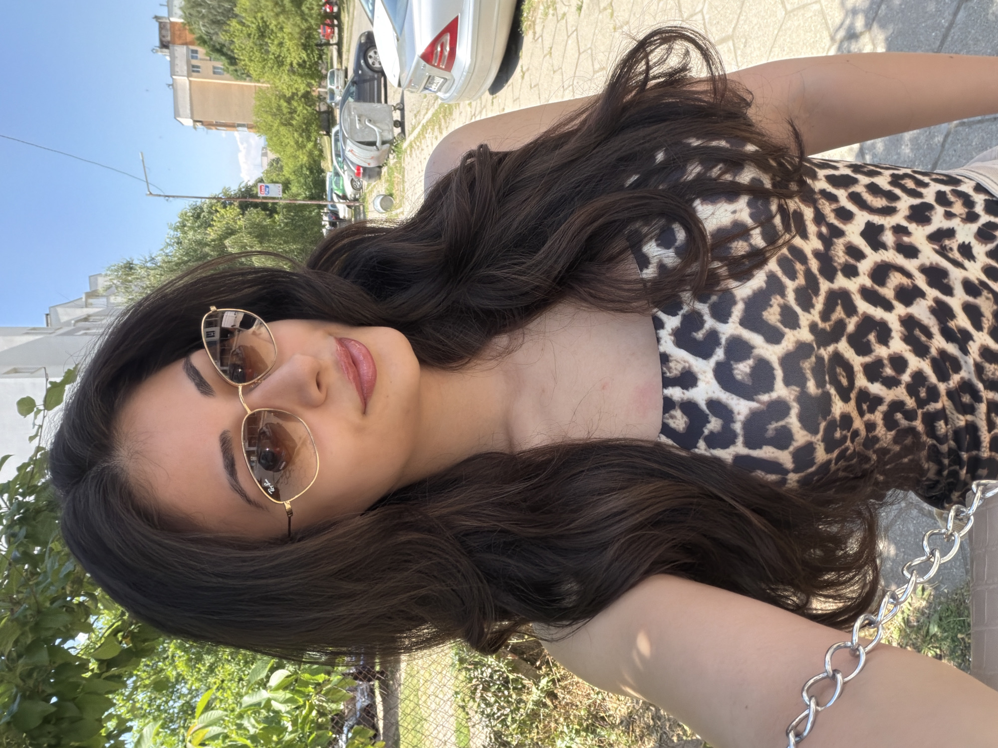A young woman is seen in a selfie, likely taken outdoors during daylight. She is wearing sunglasses with gold frames and a leopard-print top. She also has a silver chain bracelet on her wrist. The background features a street scene, including parked cars and buildings, suggesting an urban environment. Based on visual details, the location could be Blagoevgrad, Bulgaria.