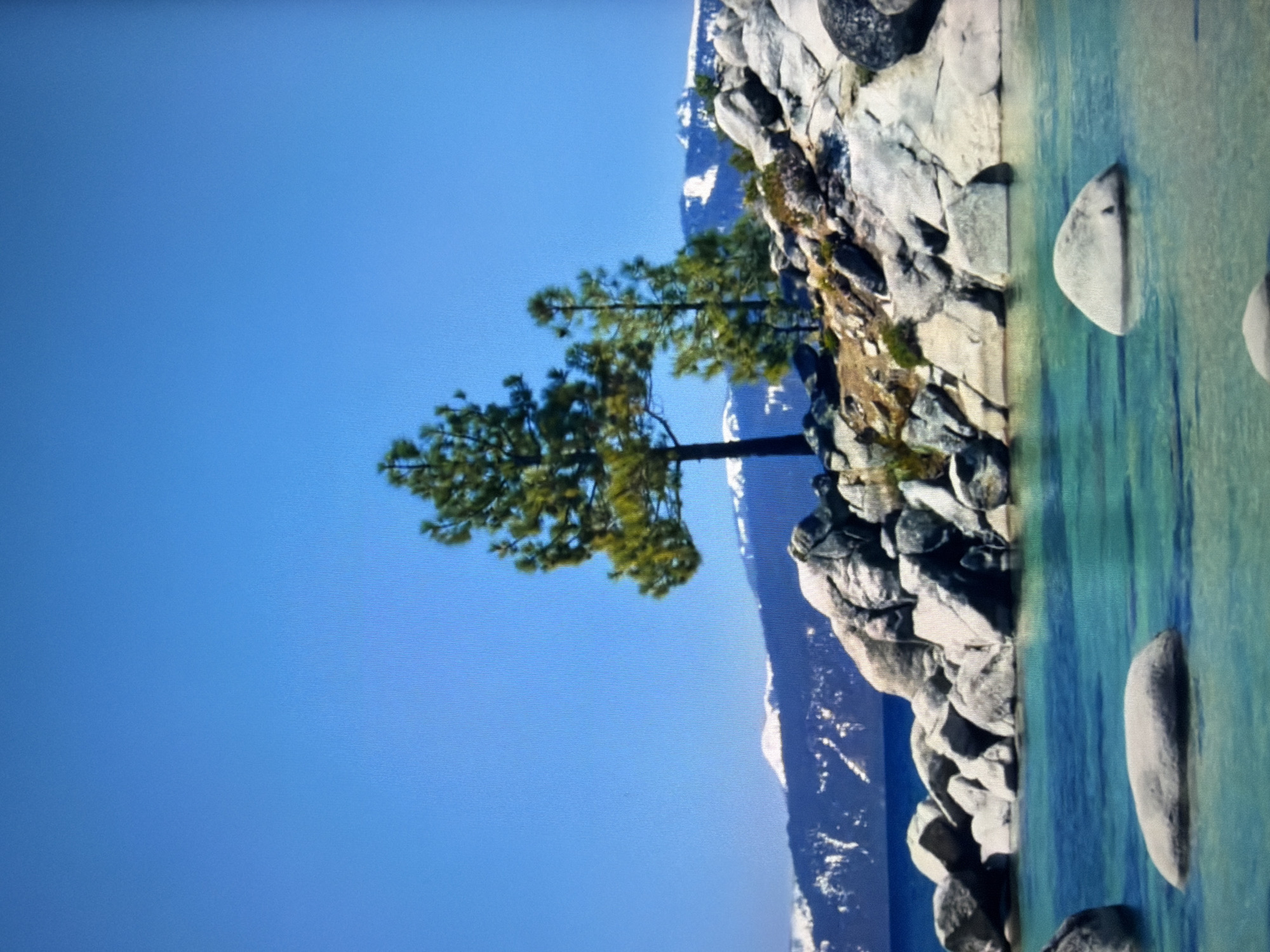 The image depicts a natural landscape with a clear blue sky above. In the foreground, turquoise water surrounds several large, smooth rocks. The middle ground features a rocky shoreline with scattered green vegetation and two evergreen trees. In the background, snow-capped mountains form a distant horizon. The scene is a serene natural environment. This image is not located in the city of Kyiv, Ukraine.
