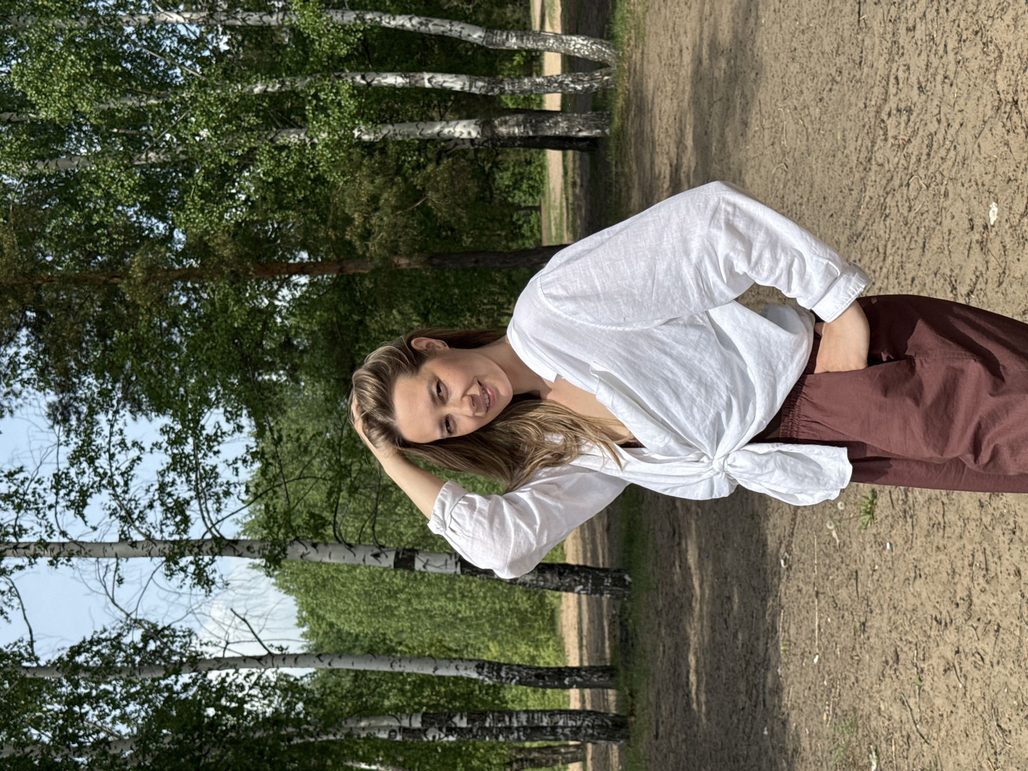 A young woman stands on a dirt path in a forest. She wears a white, long-sleeved blouse tied at the waist and brown pants. She has light brown hair and a slight smile. Her right hand is touching her hair. The background includes birch trees and green foliage. The scene is outdoors and appears to be on a sunny day in a forest in Noviy, Russia.