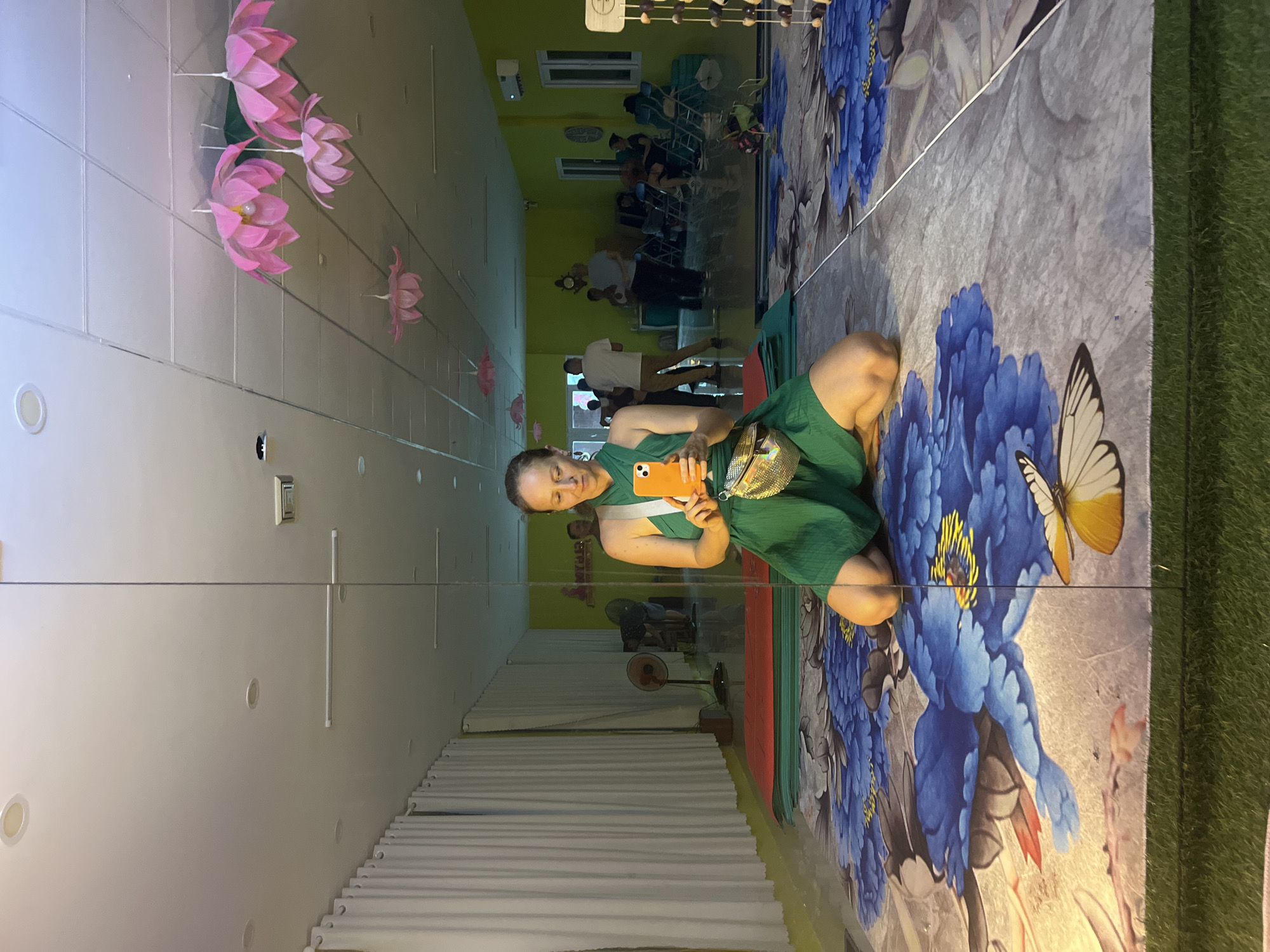 A woman is kneeling on a large, colorful yoga mat with a floral and butterfly pattern, taking a selfie in a mirrored room that appears to be a yoga studio. She is wearing a green dress and a gold fanny pack, and holding an orange smartphone. In the reflection, other people are visible in the background, some sitting and some standing, with yoga mats and fans on the floor. The room has bright green walls and a white ceiling with decorative pink lotus-shaped lights. The overall impression is a relaxed and possibly social atmosphere within a dedicated wellness space. The image was taken in a yoga studio in Đà Nẵng, Vietnam.