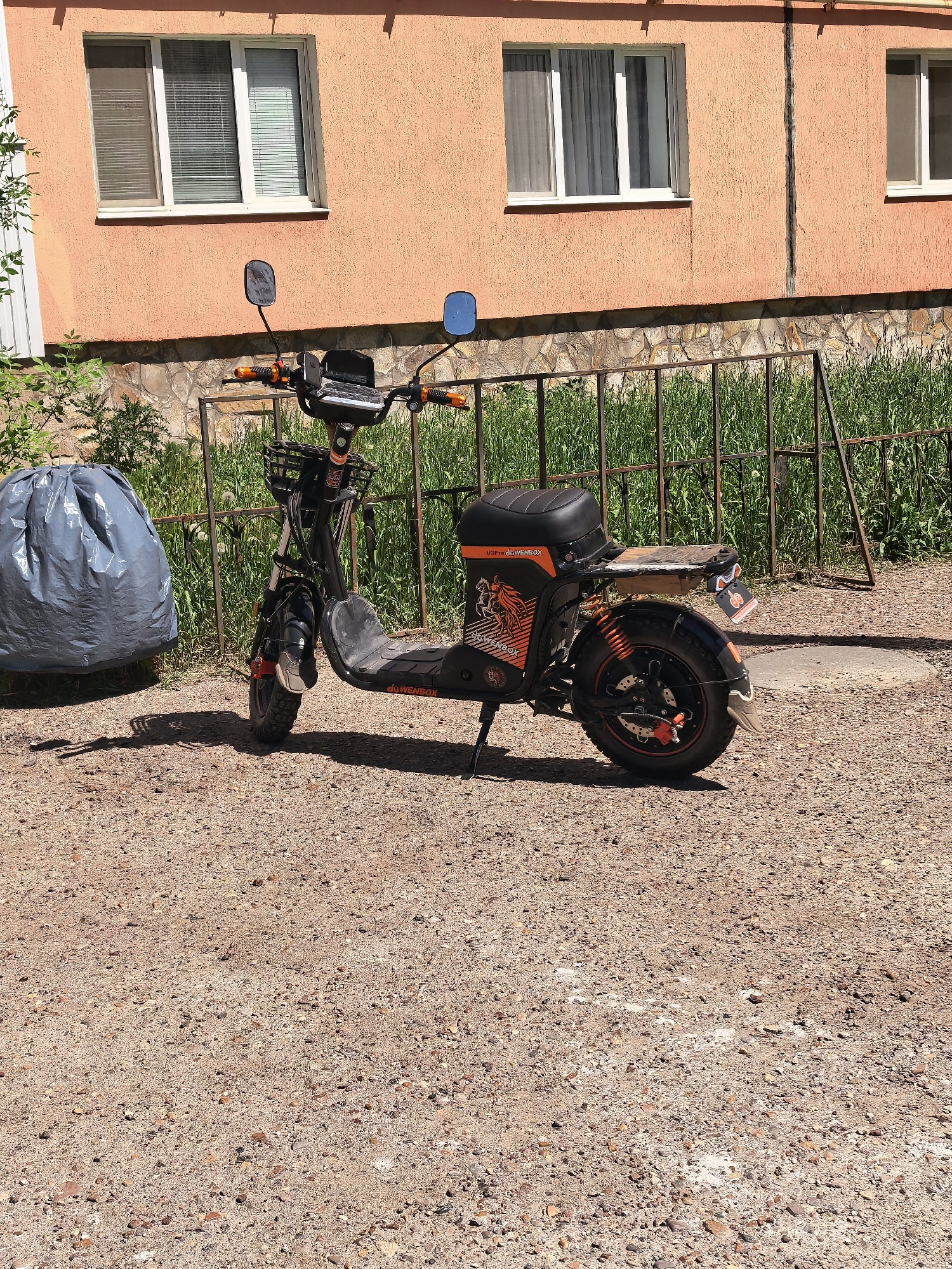 The image depicts an electric scooter parked outdoors on a dirt and gravel surface. Behind the scooter is a low, wrought-iron fence with grass growing behind it. To the left of the scooter, there is a large gray trash bag.
In the background is a peach-colored building with multiple windows. Based on the location, the scene is in Sterlitamak, Russia. There are no people visible in the image. The scooter appears to be unattended.
