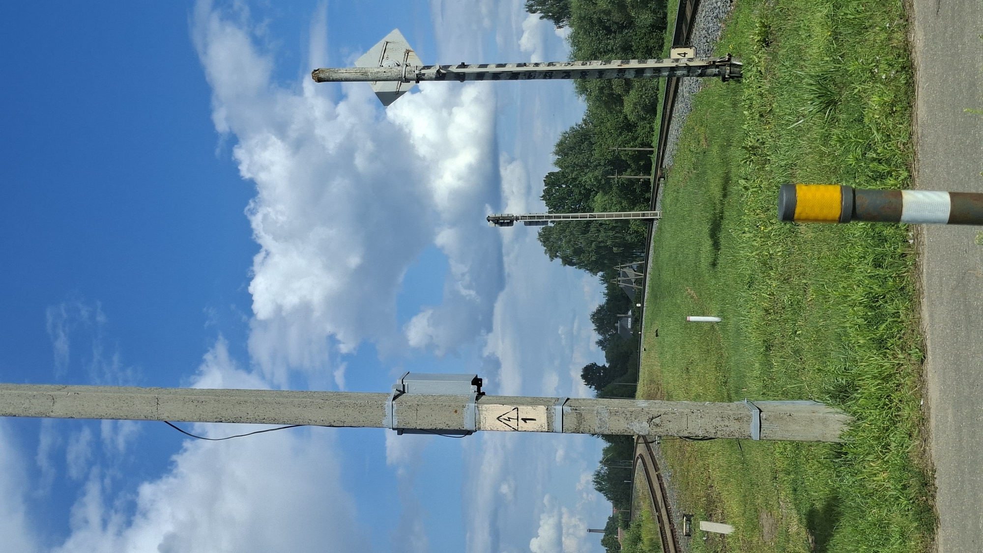 The image depicts a rural or suburban scene in Maladzyechna, Belarus, featuring railway tracks and associated infrastructure under a bright blue sky with scattered white clouds. In the foreground, a concrete utility pole stands prominently on the left, displaying a yellow and black warning sign with a lightning bolt symbol and the number '1'. A grey electrical box is attached to the pole. To the right of this pole, another taller, striped pole supports a diamond-shaped sign and what appears to be a railway signal, with a ladder attached to its side. A shorter, striped bollard with yellow and white reflective bands is positioned even further to the right and closer to the viewer. The railway tracks curve into the distance, bordered by lush green grass. Trees and some distant buildings are visible in the background. The scene appears peaceful and operational, with no people present.