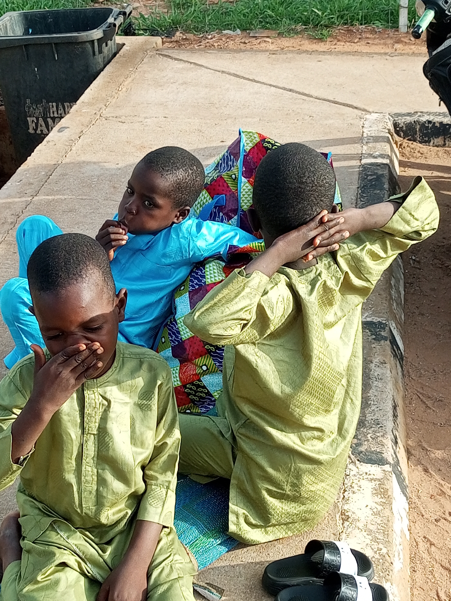 Three young boys are sitting on a concrete ledge outdoors. The boy in the foreground is wearing a light green outfit and has his right hand covering his mouth and nose. The boy in the middle is wearing a bright blue outfit and has his fingers in his mouth. The boy on the right is wearing a light green outfit and has his arms behind his head. A colorful patterned cloth is behind the two boys in the middle and right. A black trash bin is visible in the upper left corner, and grass and dirt surround the concrete ledge. Black and white sandals are placed on the ground in the lower right corner.
