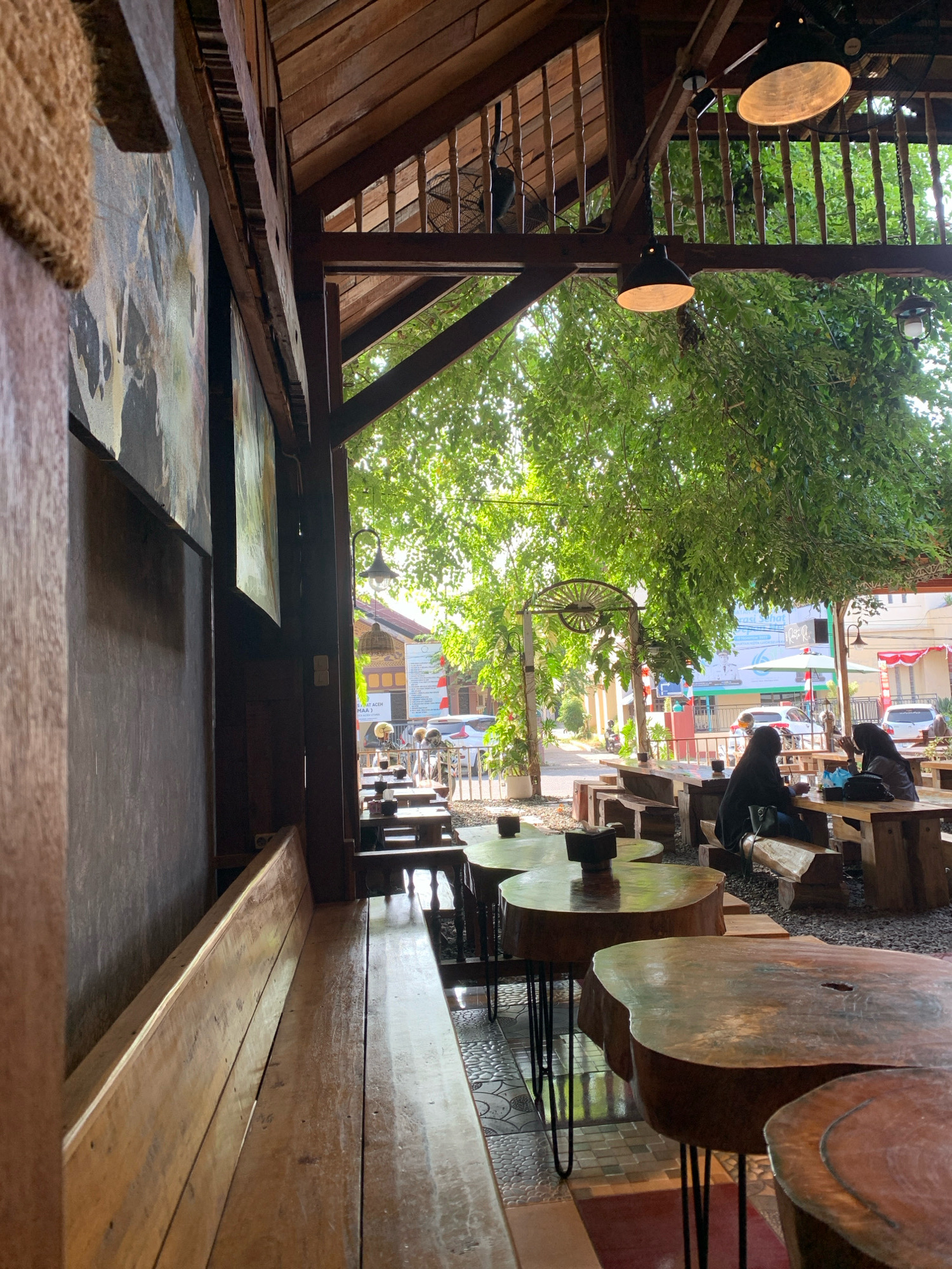 This is an outdoor seating area of a cafe, likely in Lhokseumawe, Indonesia, characterized by its rustic and natural aesthetic. The structure is predominantly made of wood, with a slatted wooden roof providing partial shade and a wooden balcony visible in the background. Hanging industrial-style lamps and a traditional-style lantern illuminate the space.
The seating area features an assortment of wooden tables with uniquely shaped, unvarnished tops, many supported by metal hairpin legs. Long, wooden benches are set up along a wall on the left side of the image. In the middle ground, several people are seated at the tables, engaged in conversation or enjoying their time at the cafe. Two individuals, women wearing headscarves, are prominently seated at one of the tables, interacting with each other. In the background, there is a street scene with vehicles and buildings, suggesting an urban or semi-urban location. Lush green trees overhang the seating area, creating a natural canopy. The ground is a mix of tiled flooring and gravel. The overall atmosphere appears to be relaxed and inviting, with a strong emphasis on natural materials and outdoor dining.