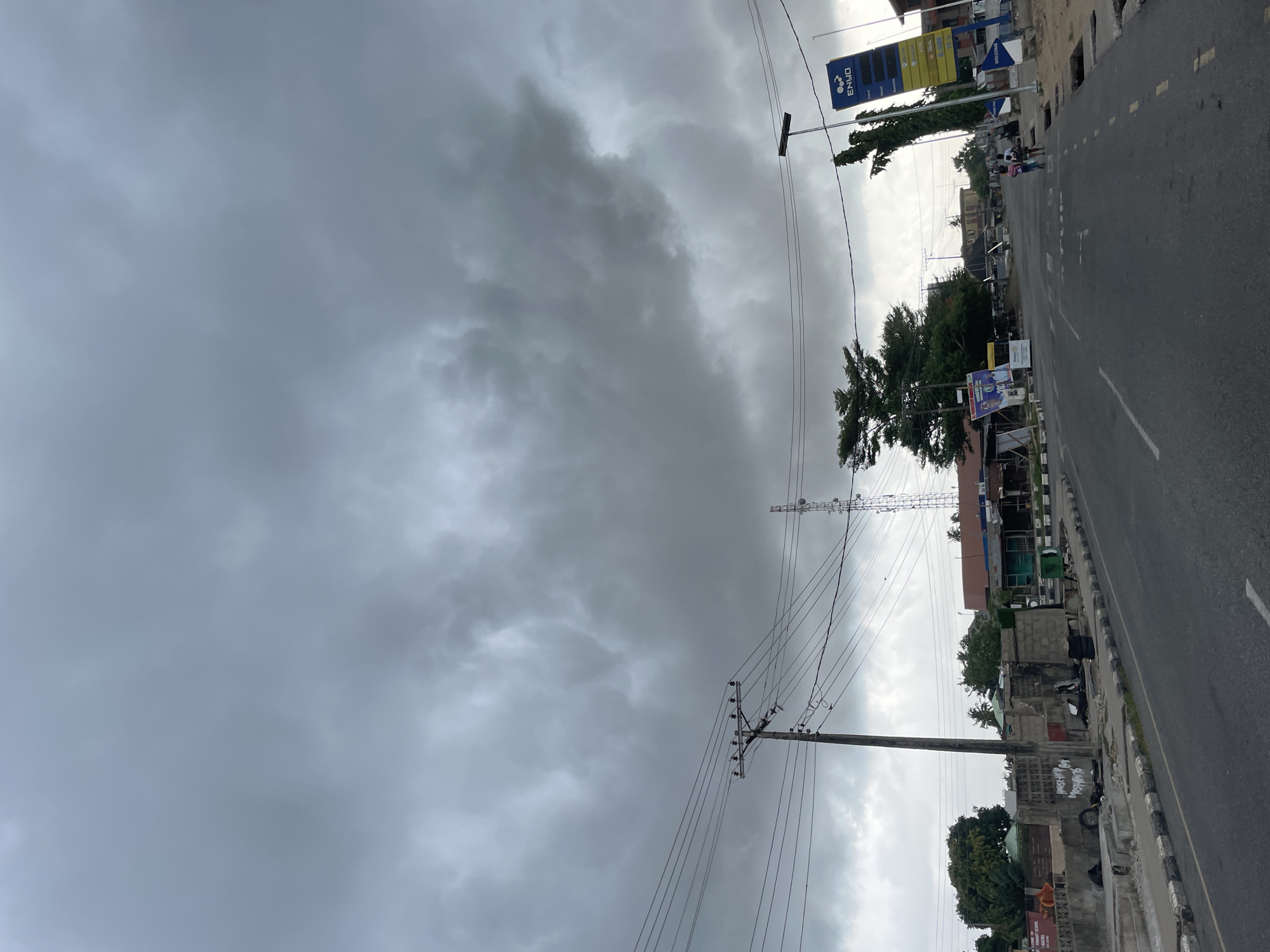 The image shows a street scene in Badagry, Nigeria, under a heavily clouded sky. A gas station with an "ENYO" sign is visible on the side of the road. Overhead power lines stretch across the view. The road itself is asphalt with white lane markings. There is a tower of some kind in the middle distance. Some people are on the sidewalk near the gas station, but are not easily visible. There are buildings and vegetation on either side of the road.