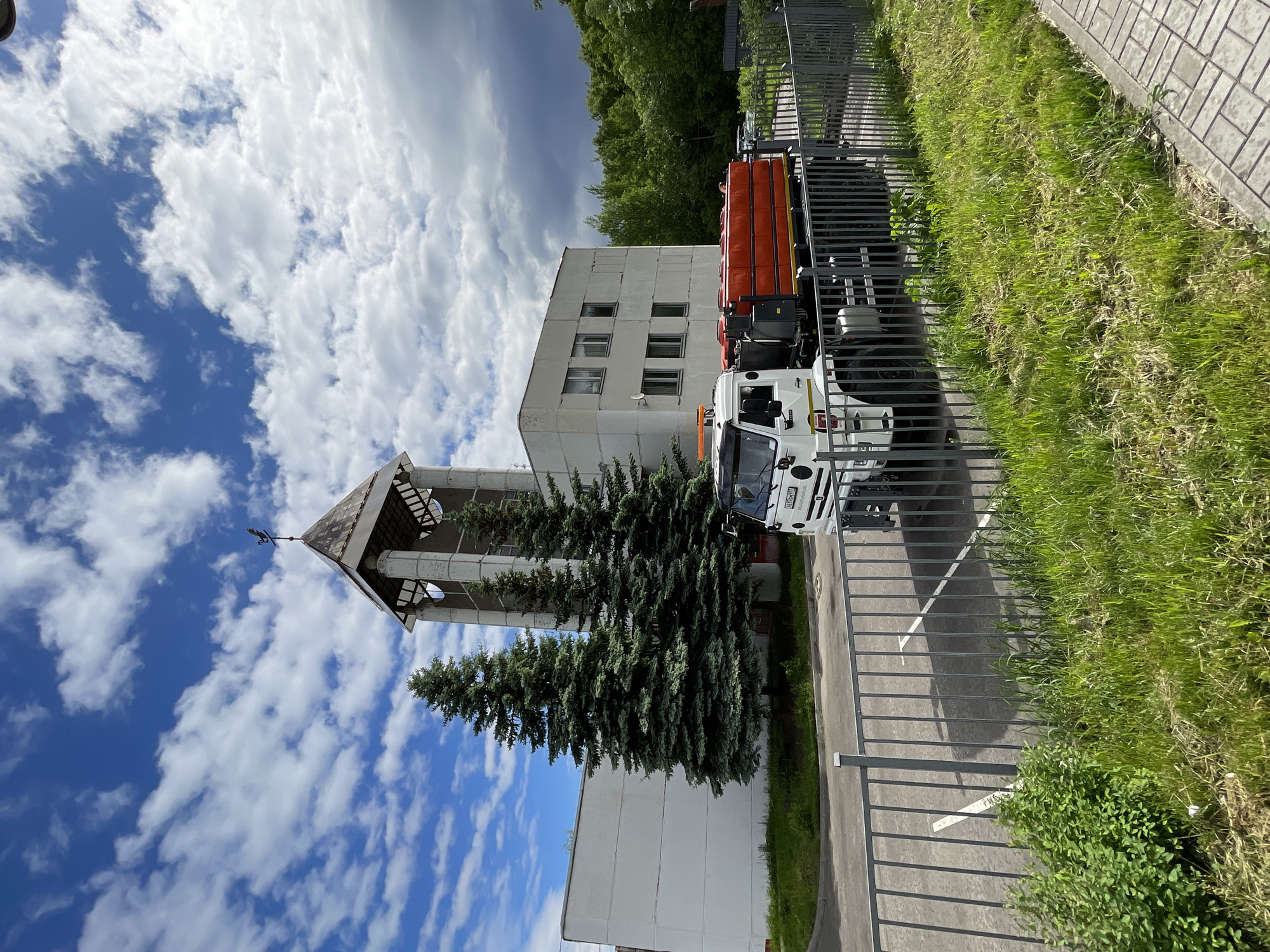 The image shows a municipal street scene in Moscow, Russia. A white and orange street-cleaning truck is parked next to a curb and a fence on the right side of the image. The truck is parked next to a building with a spire and several pine trees. The sky is blue with puffy white clouds. The environment appears to be suburban, and the scene is outdoors.