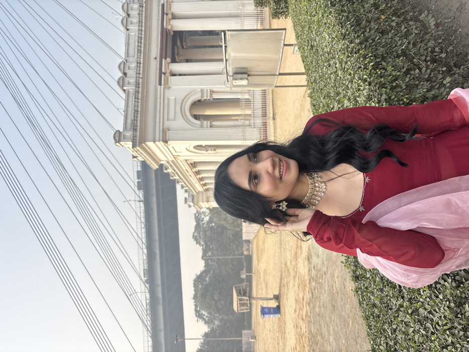 A woman in a red dress with a pale pink shawl stands outdoors. She is wearing a necklace and earrings and is smiling. Behind her are trees, a sandy area, and a white building with columns. A bridge structure with cables extends into the sky. The photo is taken at an angle, tilting the background slightly. Some bushes are visible on the right side of the frame.
