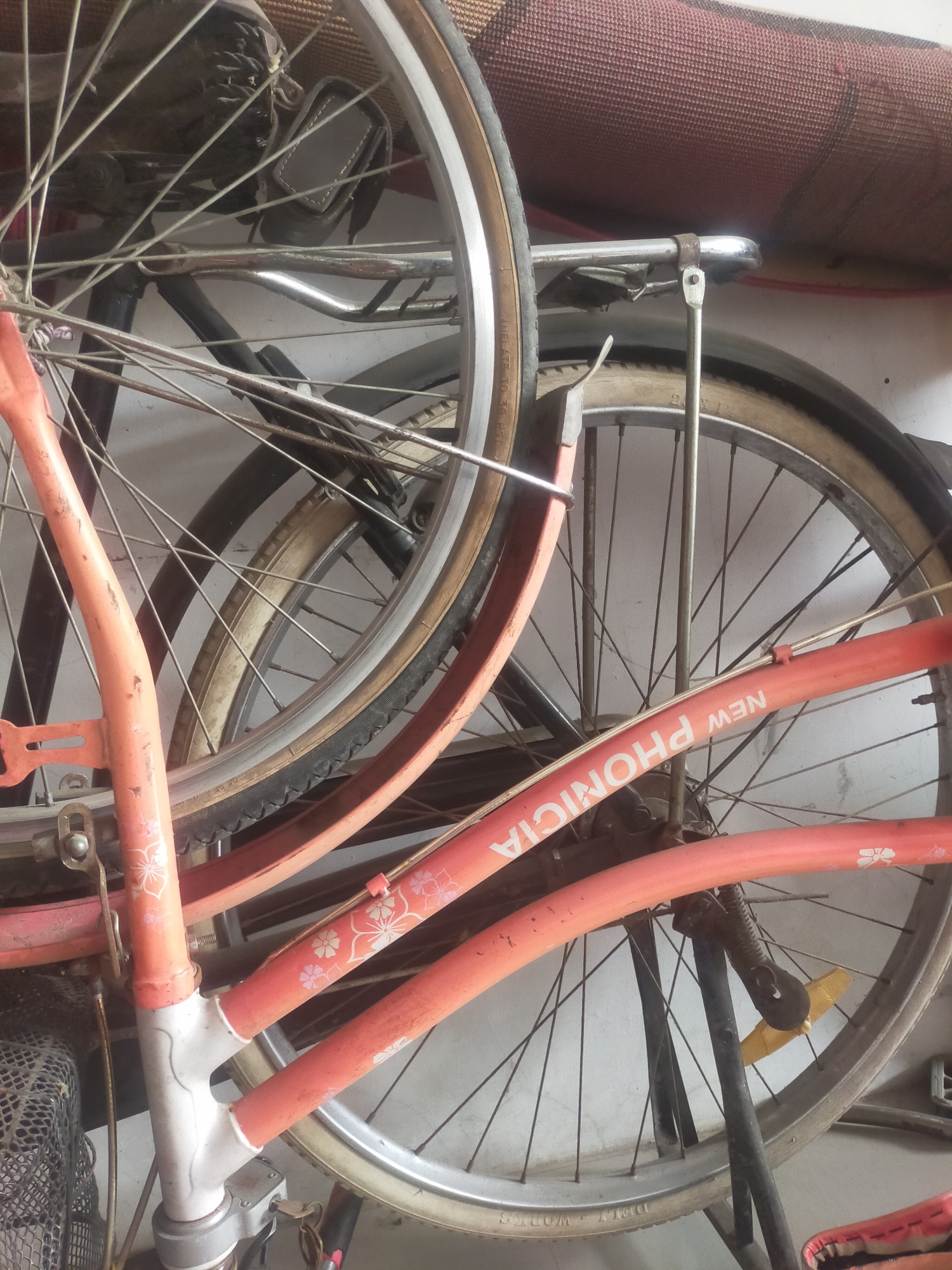 The image displays a close-up view of several bicycles stacked together, leaning against a wall. The bicycles are predominantly orange and silver. One prominent orange bicycle frame features "NEW PHONICIA" printed in white letters. Decorative floral patterns in pink and white are visible on the orange frame. Multiple wheels, spokes, and tires are visible, with some tires showing text such as "INFLATE TO 36 PSI". A rolled-up rug or mat is positioned in the background, near the top of the image. The scene appears to be an indoor storage area, possibly a garage or shed. The specific location in Tegalandong, Indonesia cannot be determined from the visual information provided in the image. There are no people visible in the image.