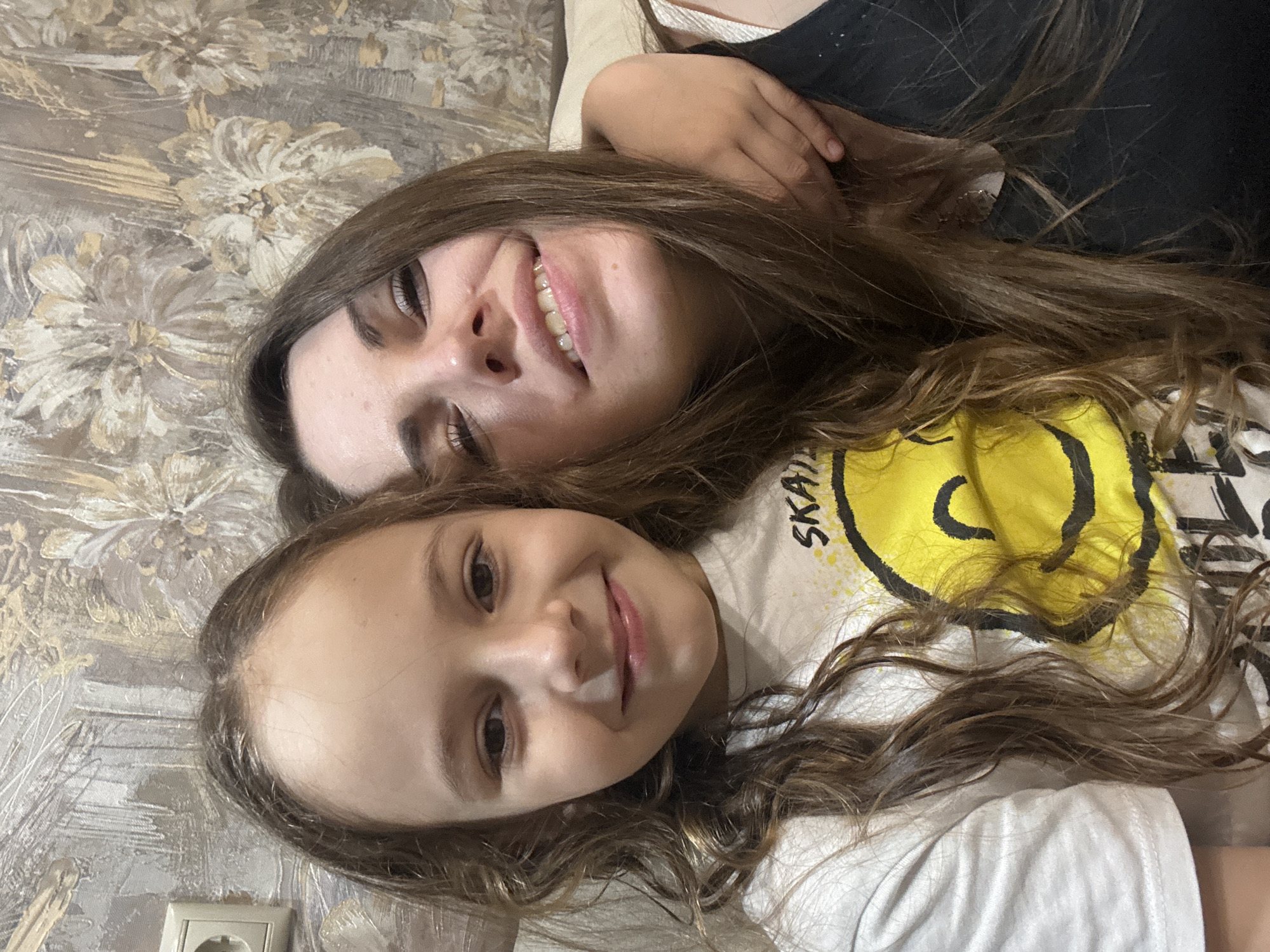 Two females, one adult and one child, are lying closely together, presumably indoors. The adult has long brown hair, a visible smile, and is wearing a black top. The child, also with long brown hair, is wearing a white t-shirt with a yellow smiley face graphic and the word "SKATE" visible. They are lying on a patterned surface resembling a bed or couch. A power outlet is partially visible in the lower left corner. The location appears to be a private residence in Раздолье, Russia.