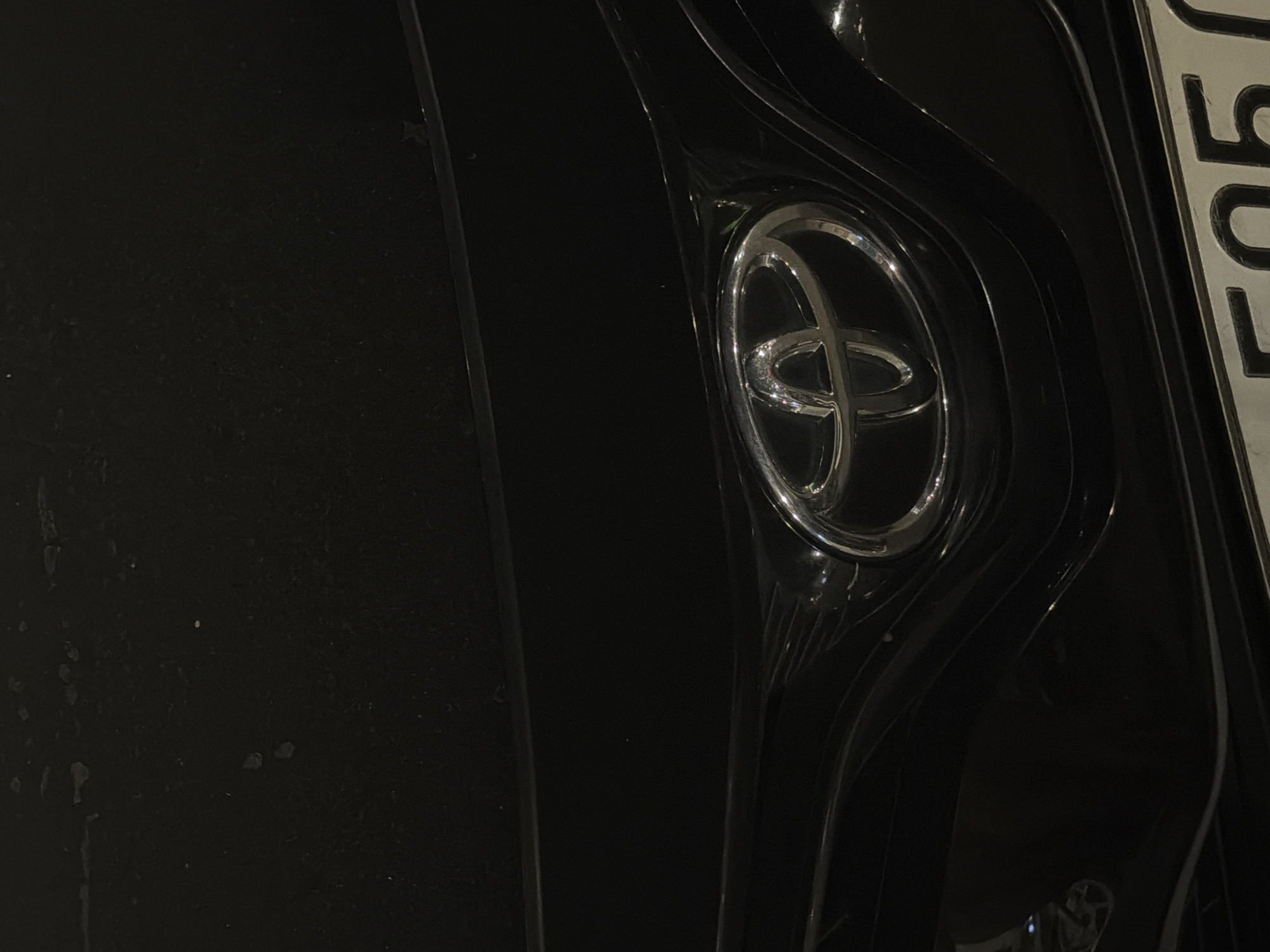 The image shows the front grille and logo of a black Toyota vehicle. The logo is a silver oval with three smaller ovals inside. The grille has a glossy black finish with some surface imperfections visible, such as small scratches and dust particles. Below the grille, a portion of a license plate is visible, with the characters "F05" partially obscured. The lighting suggests it is nighttime or a dimly lit environment. The scene is likely a street or parking area in Đà Nẵng, Vietnam, based on the context provided. There are no people or animals in the image.